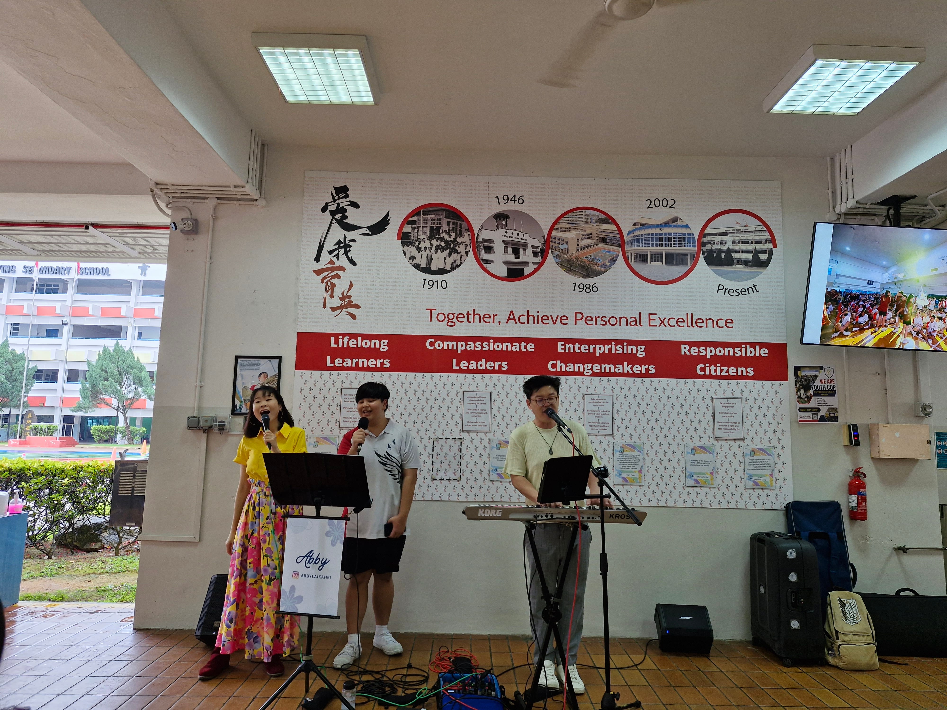 Three people sing into microphones; one plays keyboard, in front of a school wall display with photos and dates.