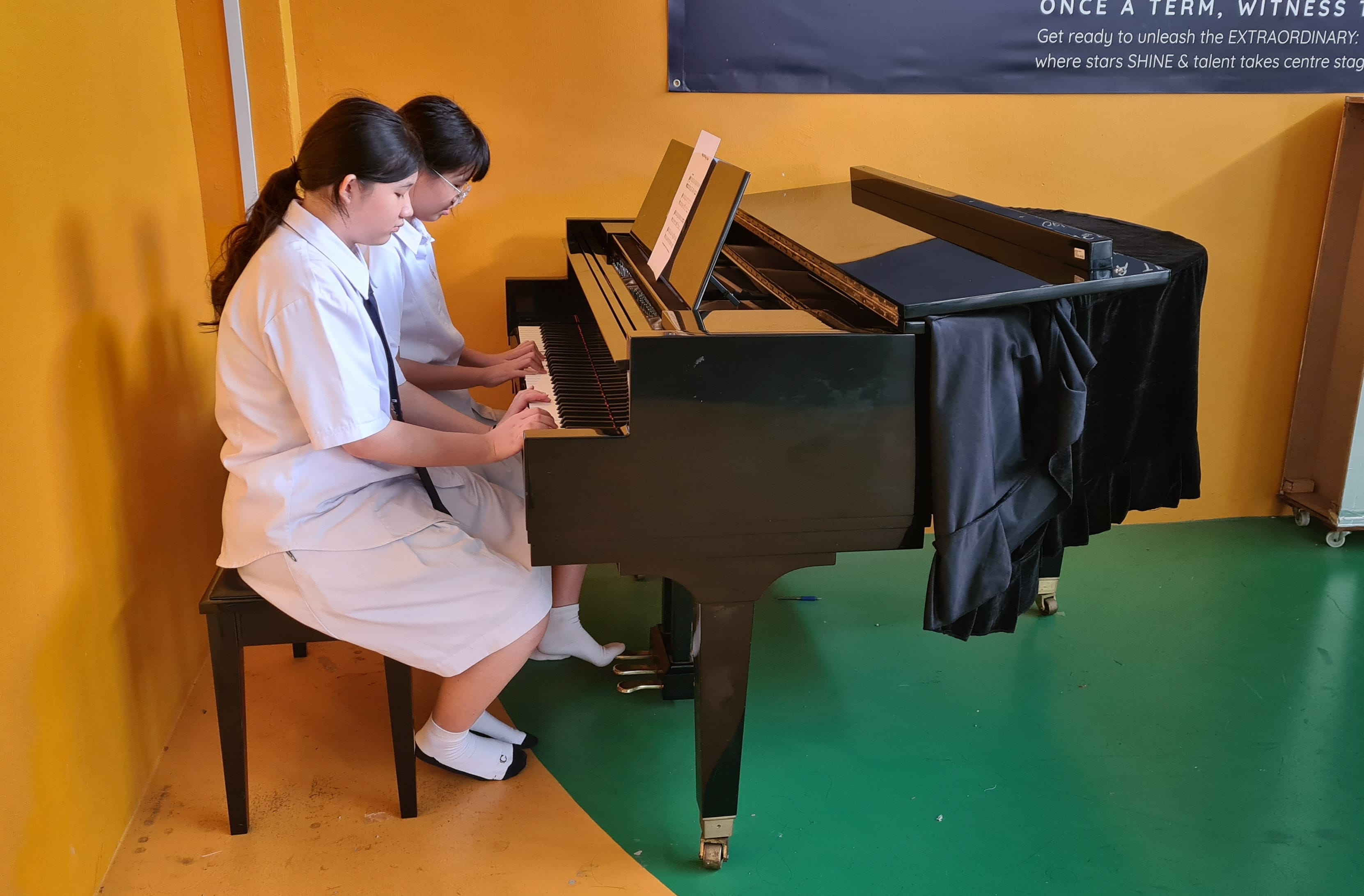 Two girls in white uniforms sit together playing a black grand piano against a yellow wall.