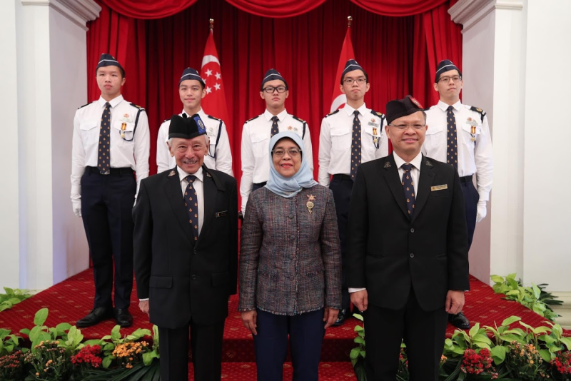 Three adults pose in front of five young uniformed cadets standing at attention before red curtains and Singapore flags.