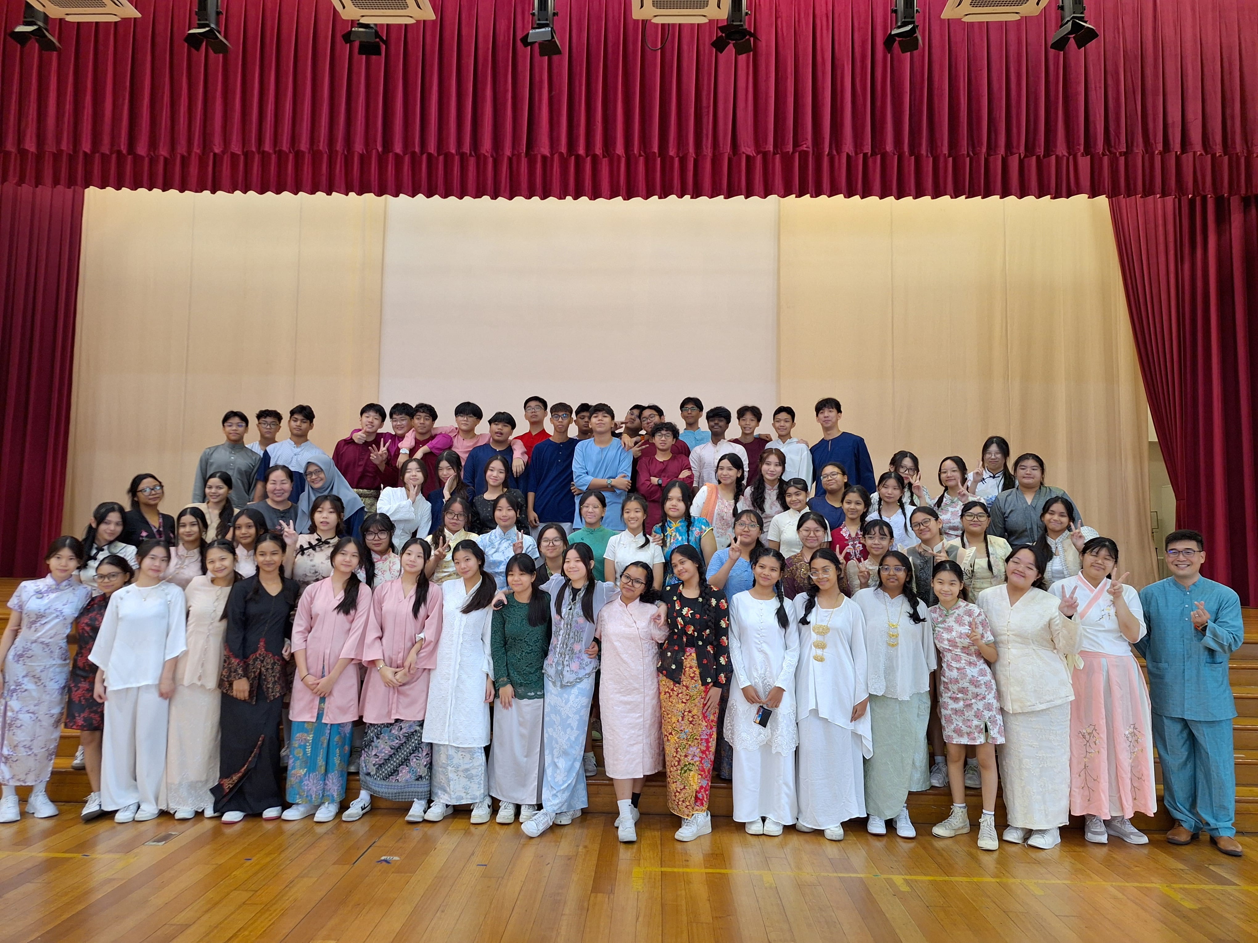 Large group of students posing on a stage under red curtains, many in colorful traditional outfits.