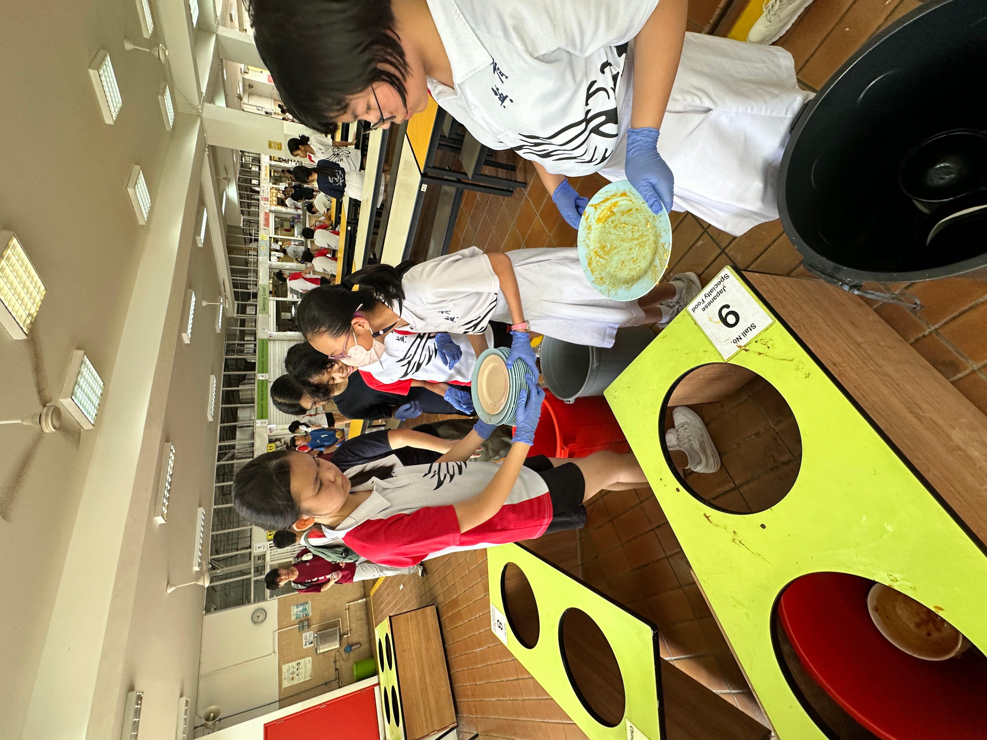 Students wearing gloves sort used plates into bins at a school canteen tray return station.