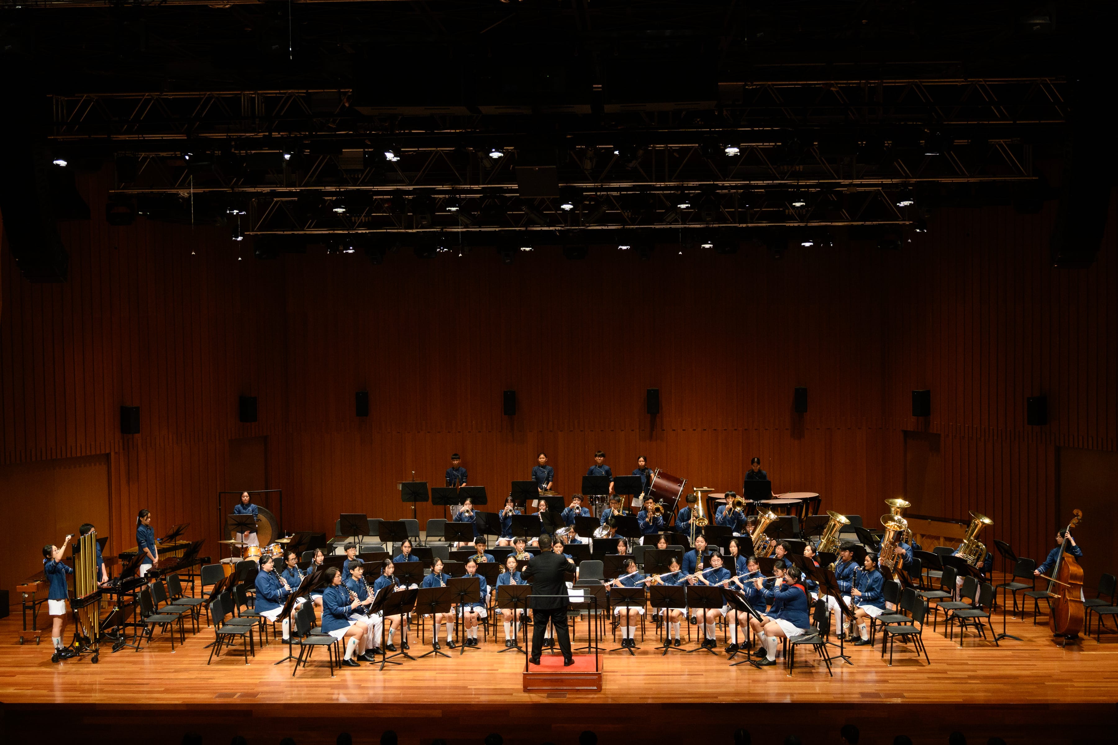 Conductor leads a seated concert band on a large wooden stage with many music stands and instruments.