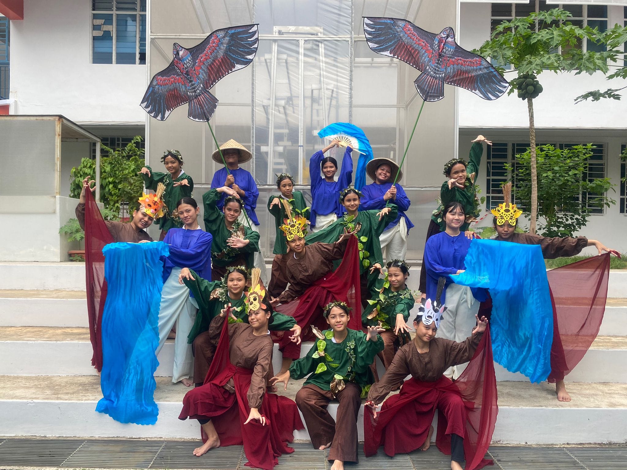 A group of children in colorful nature-themed costumes pose with bird kites, fabric, and animal masks outdoors.