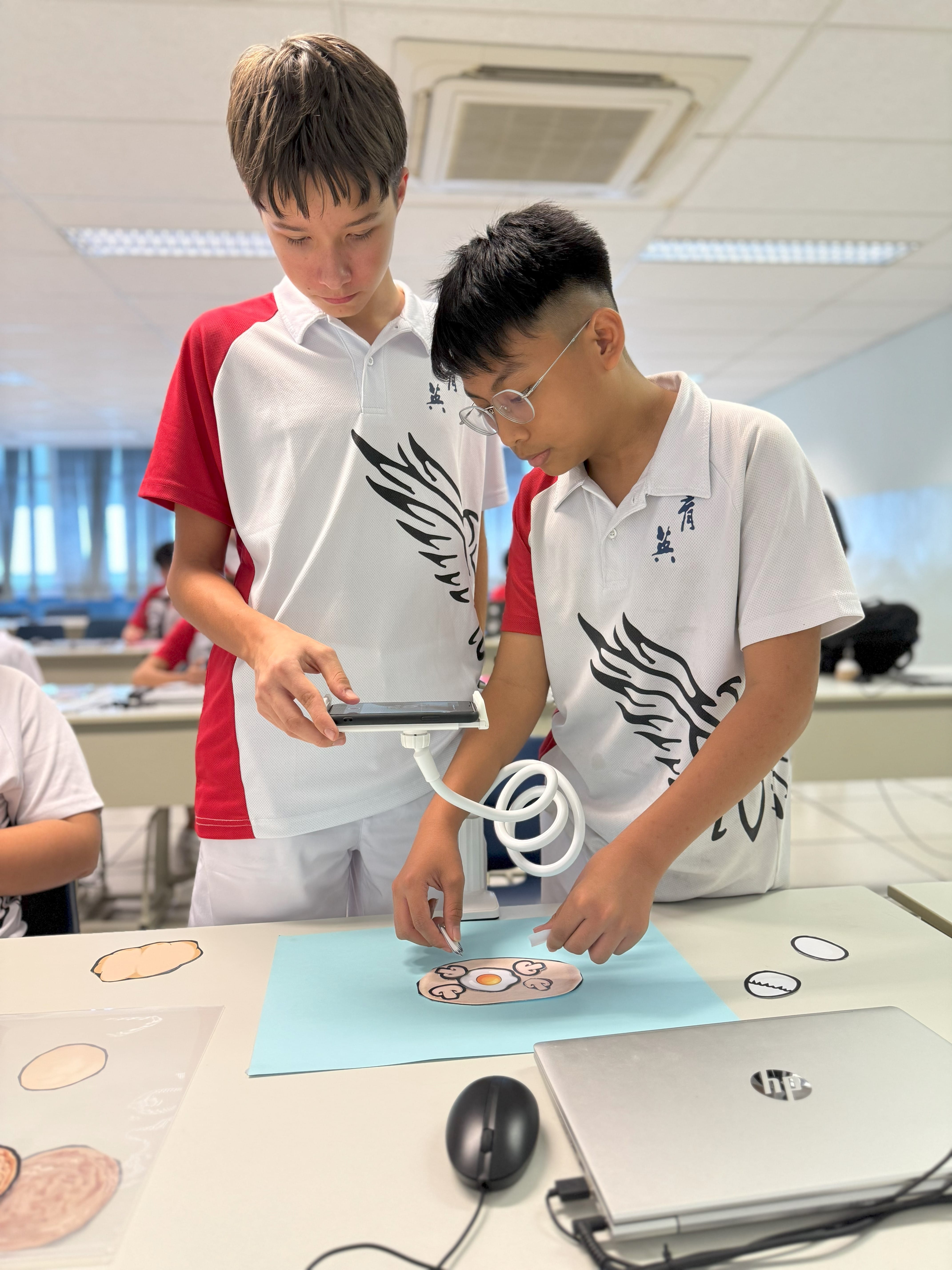 Two boys in matching white uniforms work at a desk, one holding a phone over paper cutouts near a laptop.