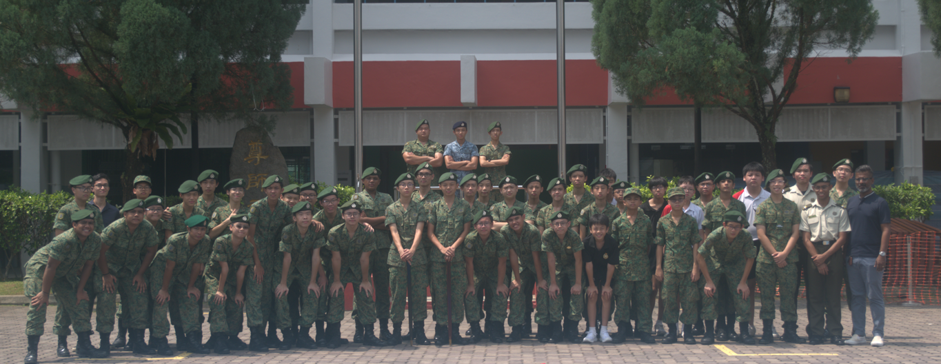 Large group of soldiers in green camouflage uniforms and berets posing outdoors in front of a red and white building.