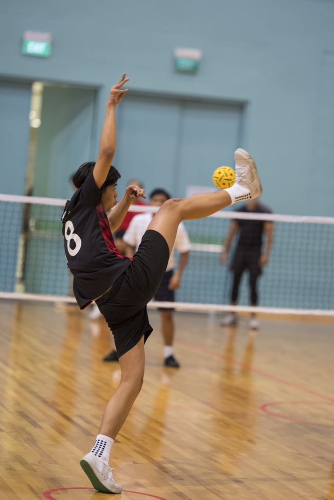 Player in black jersey number 8 kicks a yellow ball mid-air on an indoor sepak takraw court.