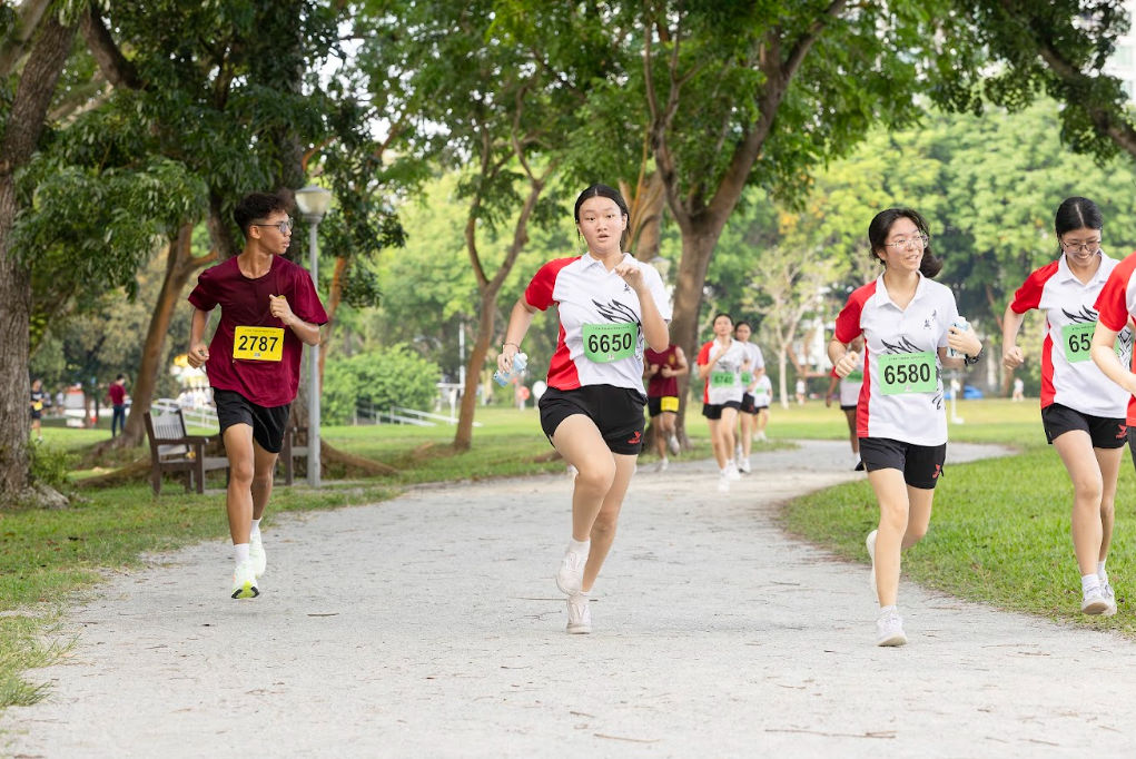 Several young runners wearing numbered bibs race along a tree-lined park path during an outdoor race event.