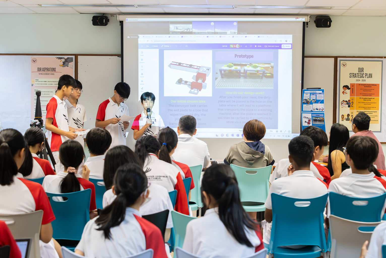 Three students present a prototype project to seated classmates in a school classroom with a projected slideshow.