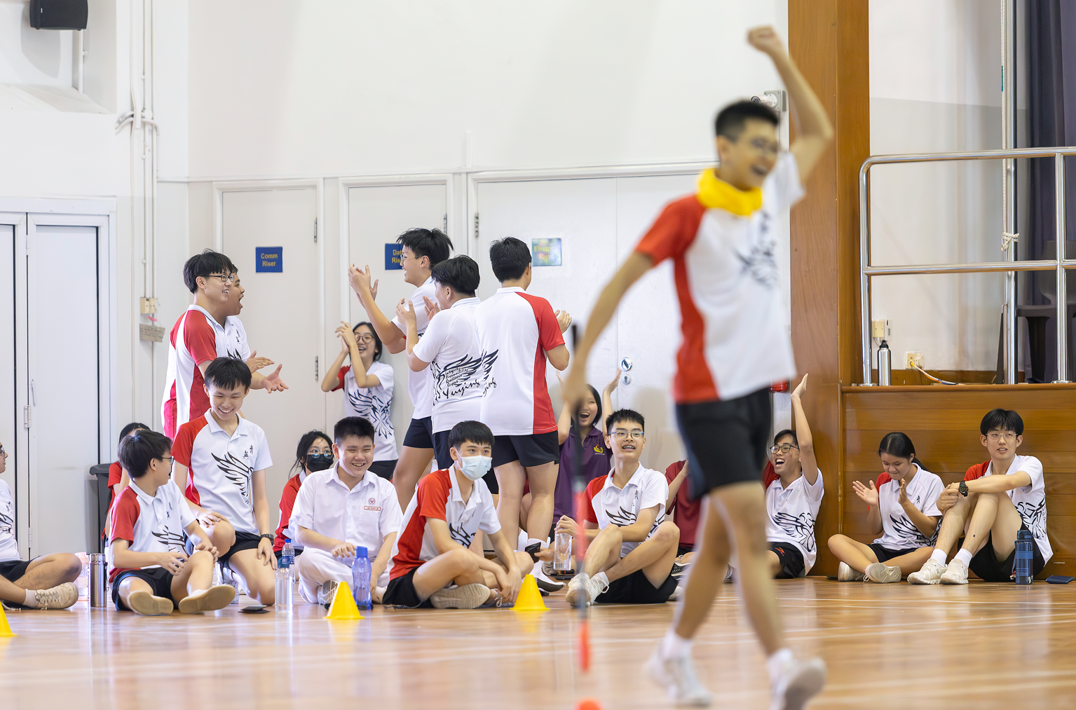 Students in white and red uniforms cheer on a gymnasium floor as a boy raises his fist in celebration.