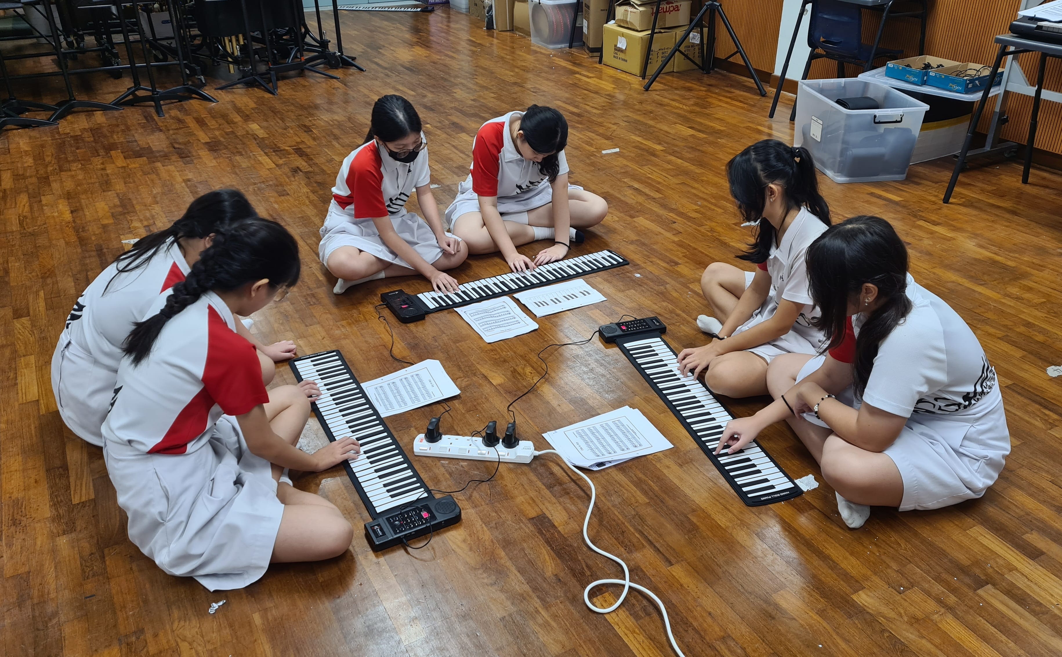 Five uniformed students sit cross-legged on a wooden floor playing three roll-up piano keyboards with sheet music.
