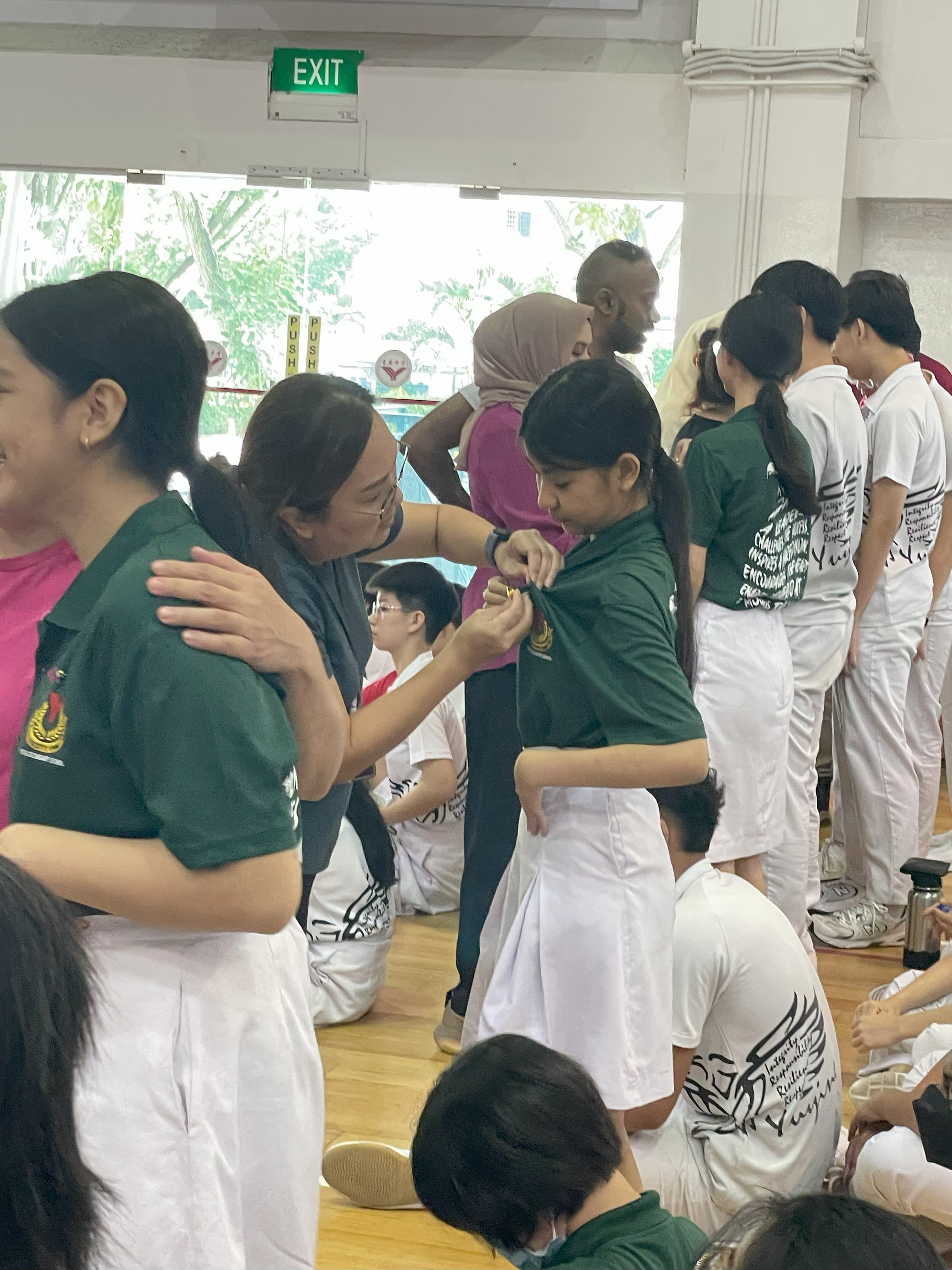 An adult pins a medal on a student in a green shirt during an indoor school ceremony with other students nearby.