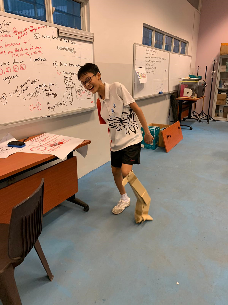 A laughing boy in a classroom wears a cardboard prosthetic leg strapped to his knee, standing near a whiteboard.