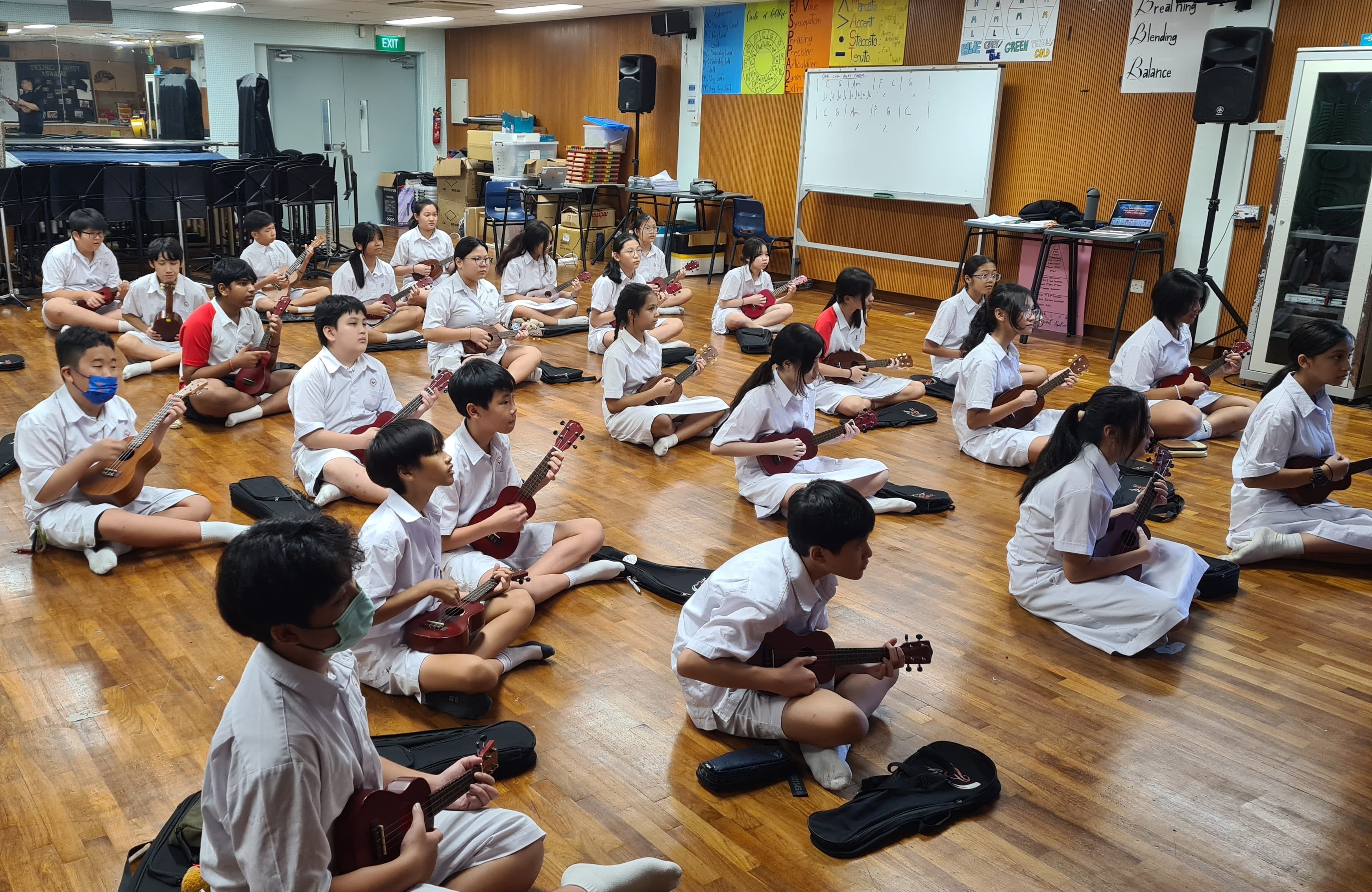Students in white uniforms sit on a wooden floor in a music classroom, each holding a ukulele.