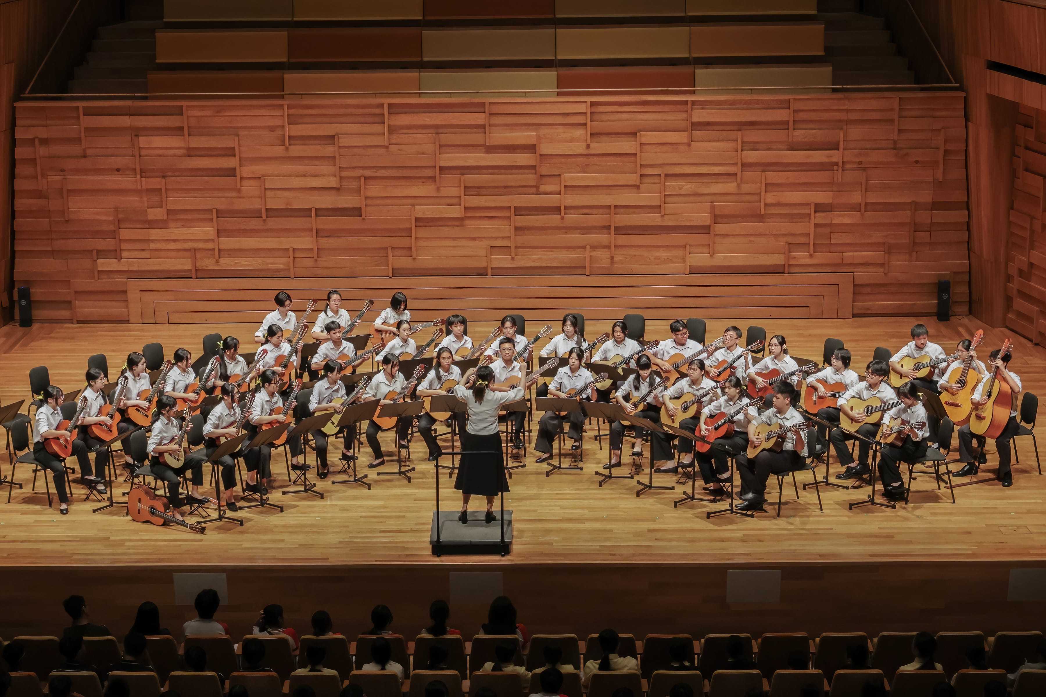 A large youth guitar ensemble in white shirts performs on a wooden concert stage, led by a standing conductor.