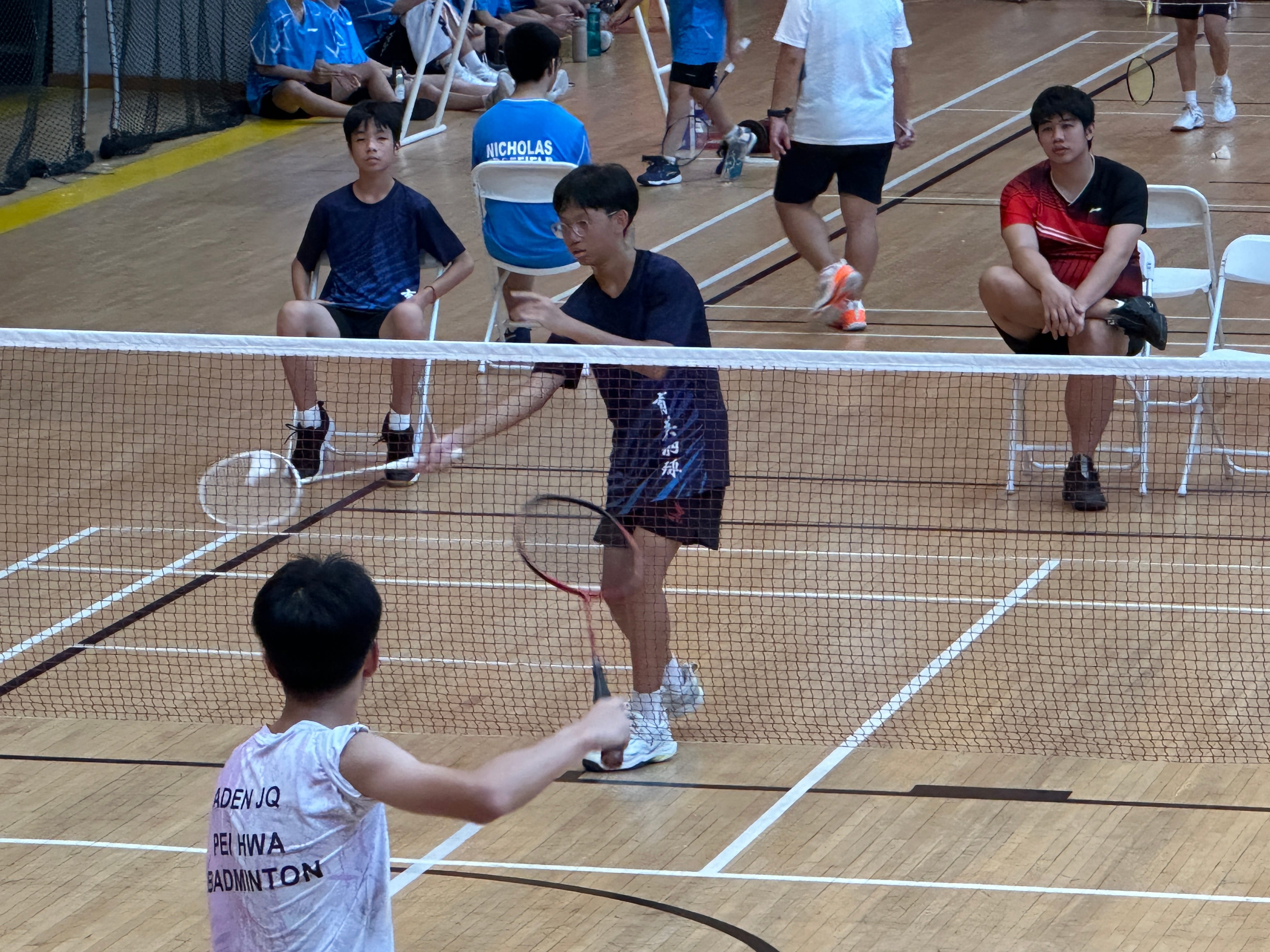 Two young men playing badminton at a net on an indoor court while others watch from chairs nearby.
