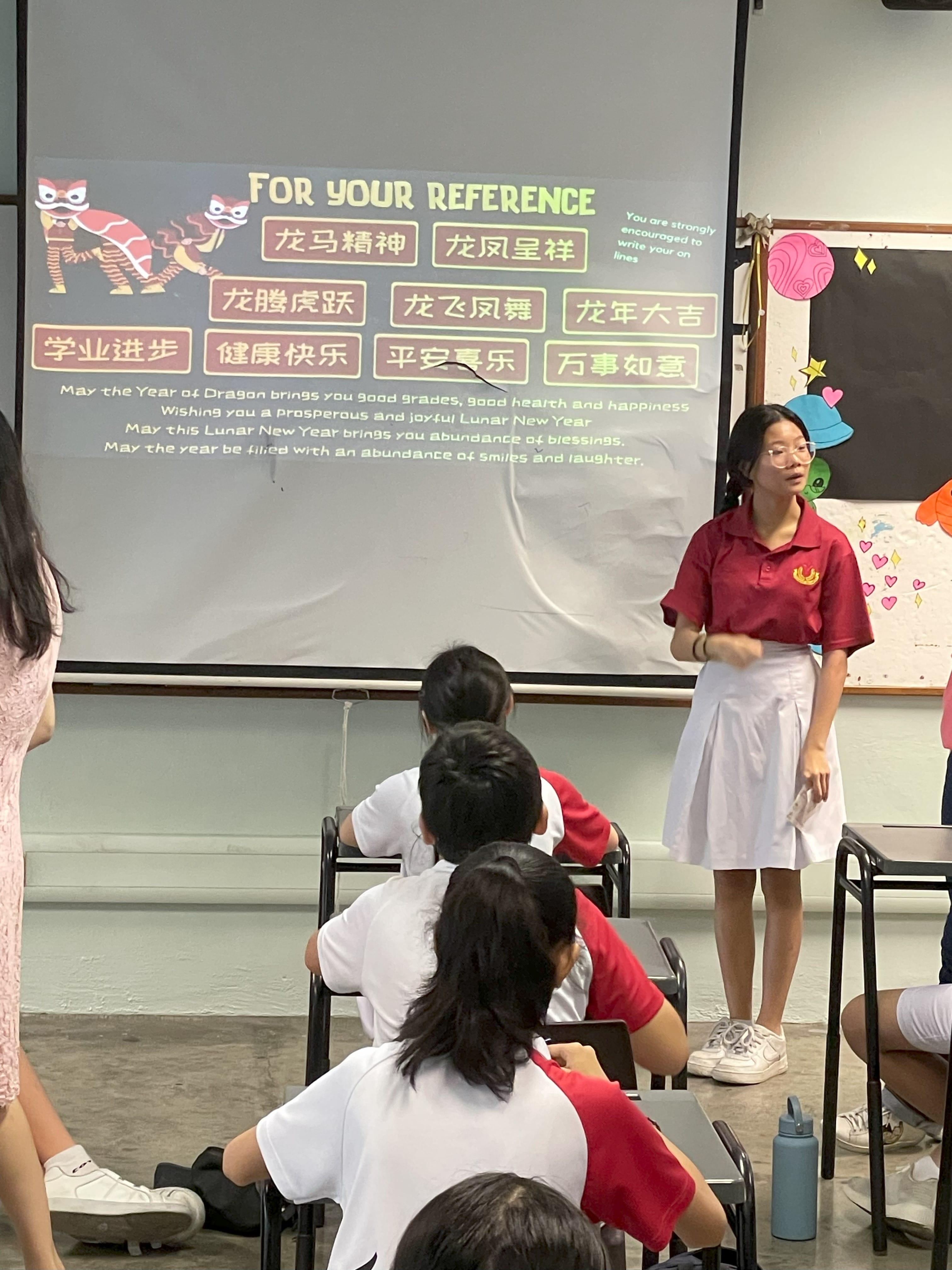 A student in a red uniform presents a Lunar New Year slideshow with Chinese phrases to seated classmates in a classroom.