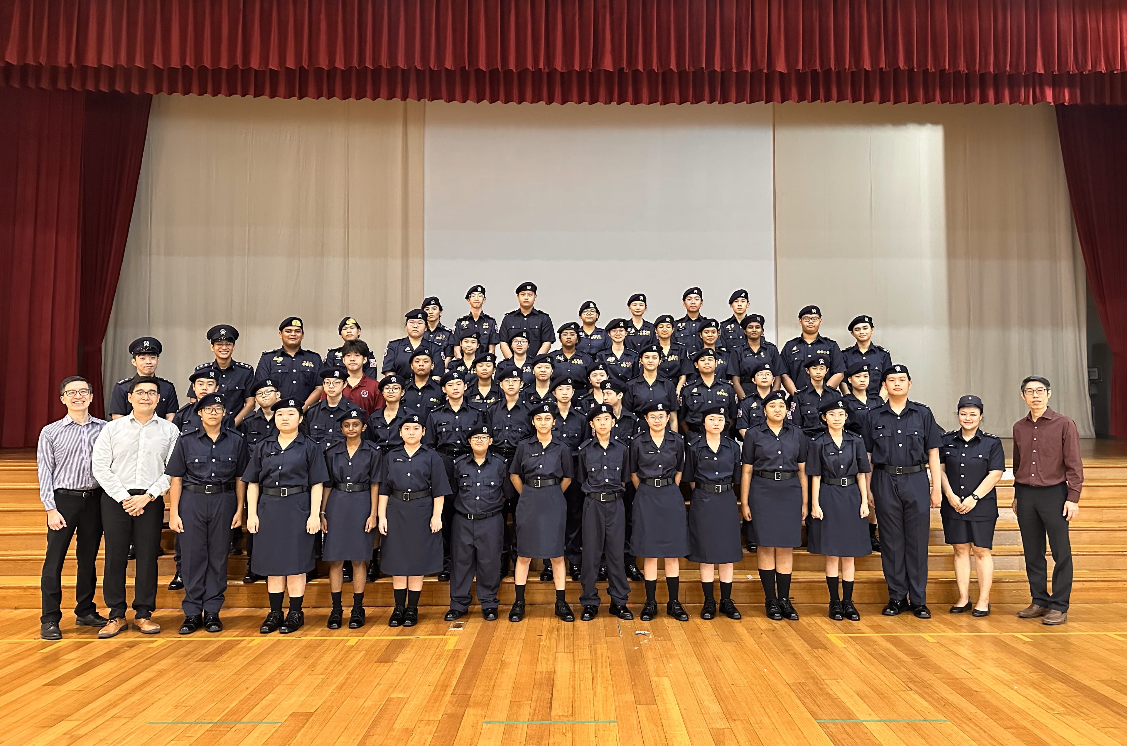 Large group of youth in dark navy uniforms and berets posing on a wooden stage floor with red curtains behind.