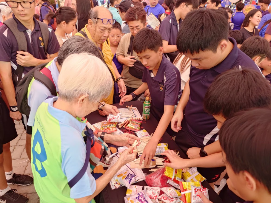 A crowd of people gather around a table covered with snack packets and small craft items at an outdoor event.