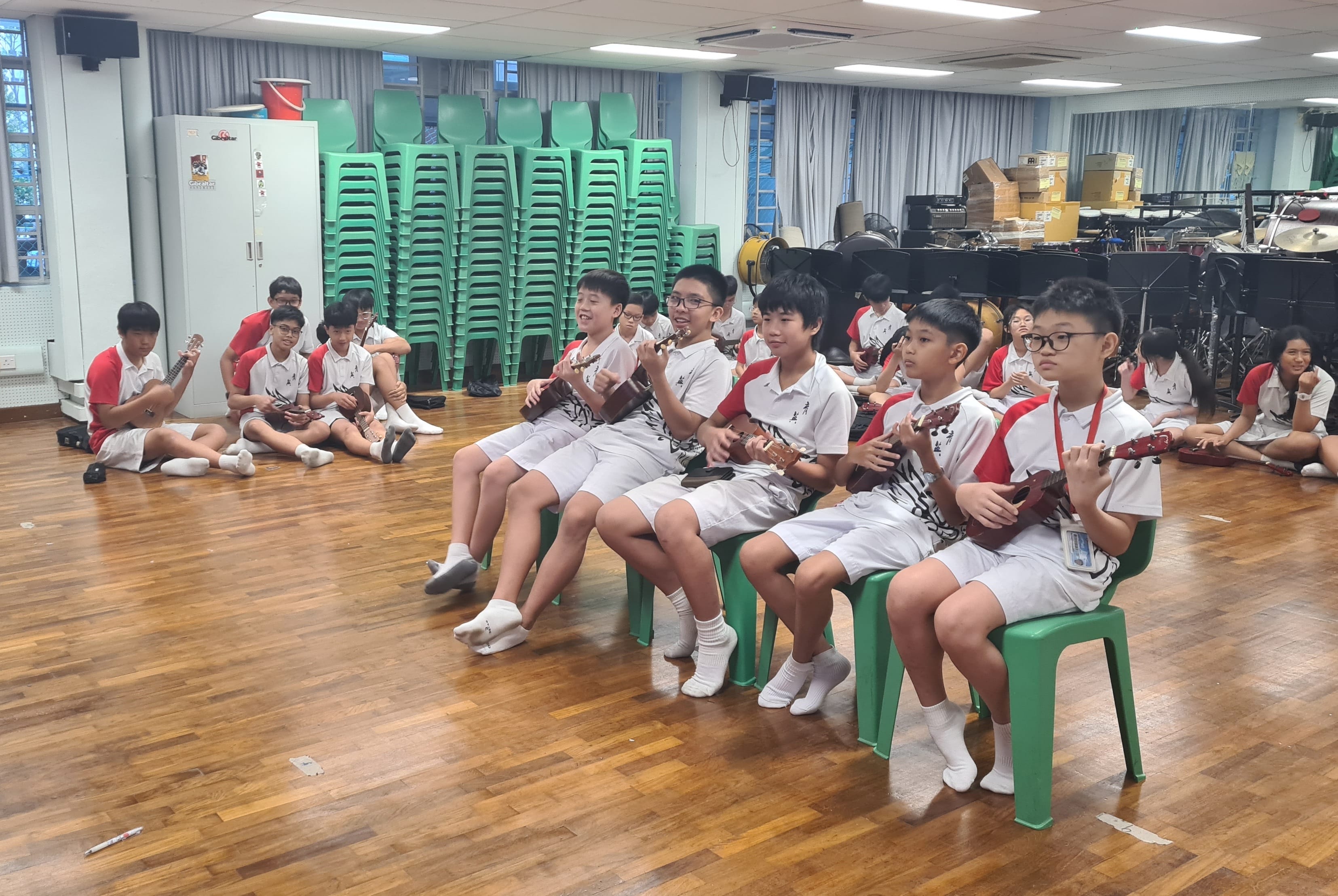 Students in white and red uniforms sit on green chairs and the floor playing ukuleles in a school music room.