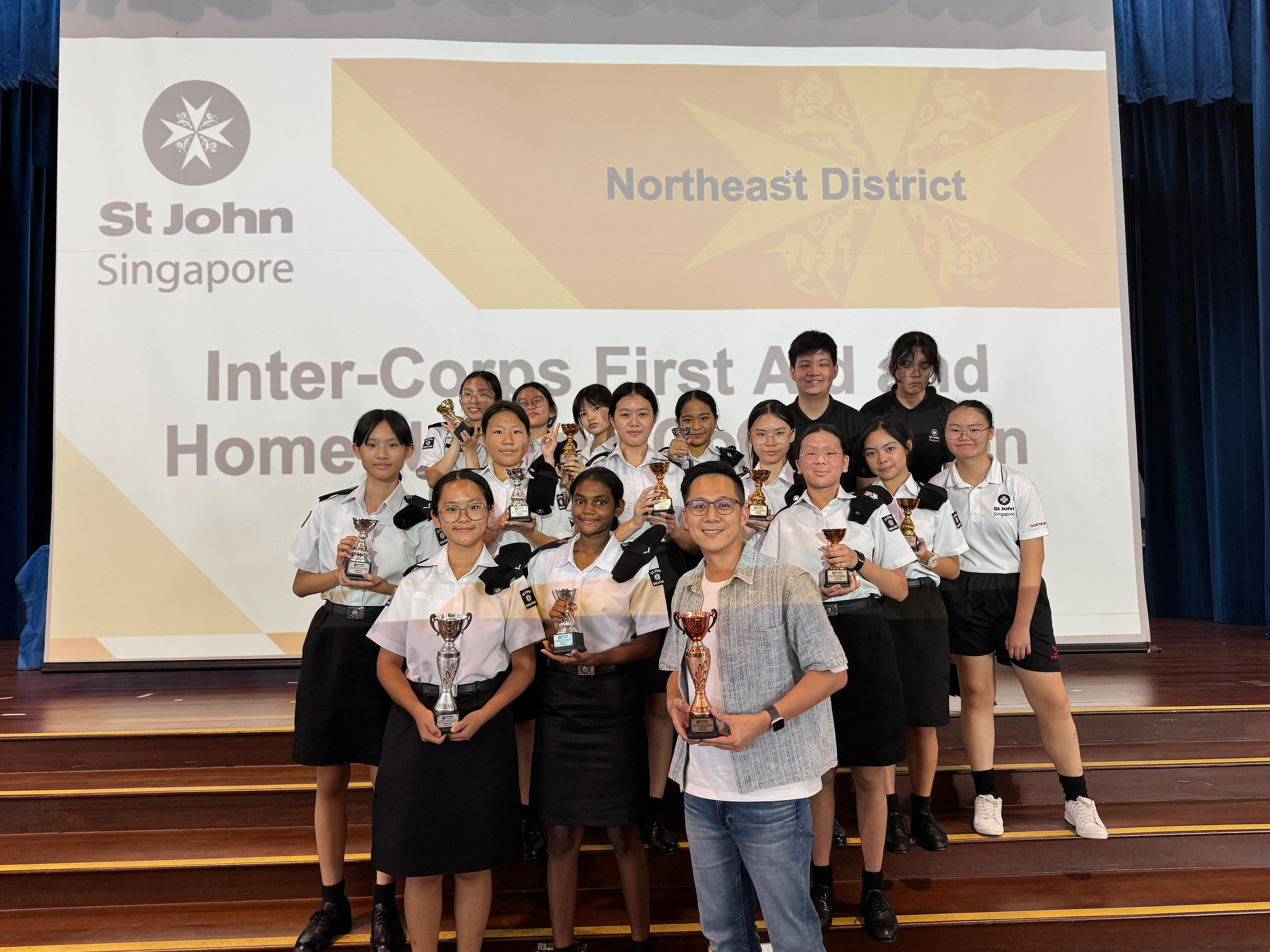 Group of St John Singapore cadets and an adult holding trophies in front of an Inter-Corps First Aid competition backdrop.