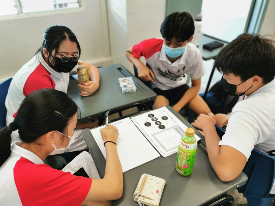 Four masked students in school uniforms sit around a desk, working together on papers and a notebook.