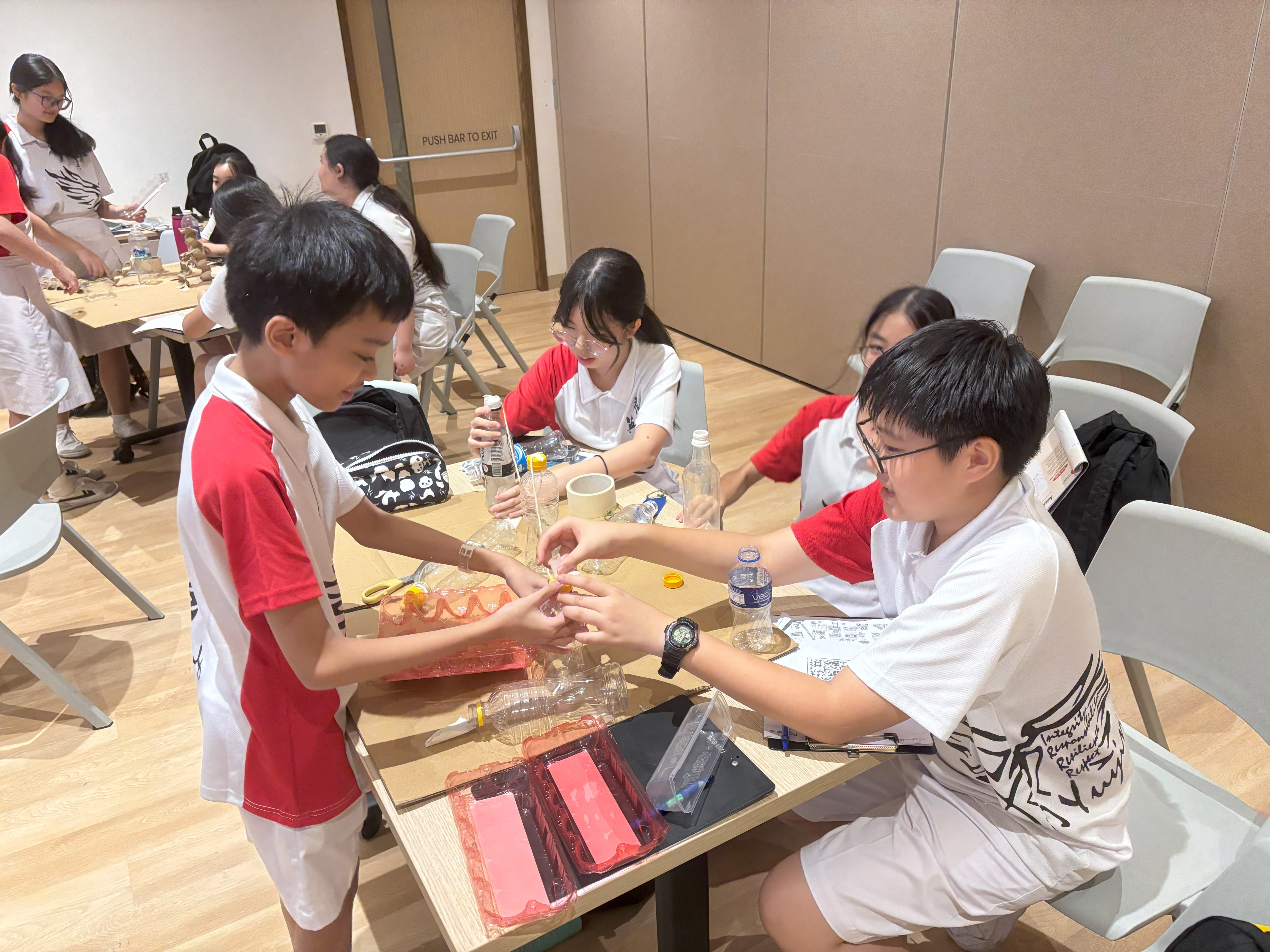 Students in red-and-white shirts build a project at tables with bottles, cups, and tools in a classroom.