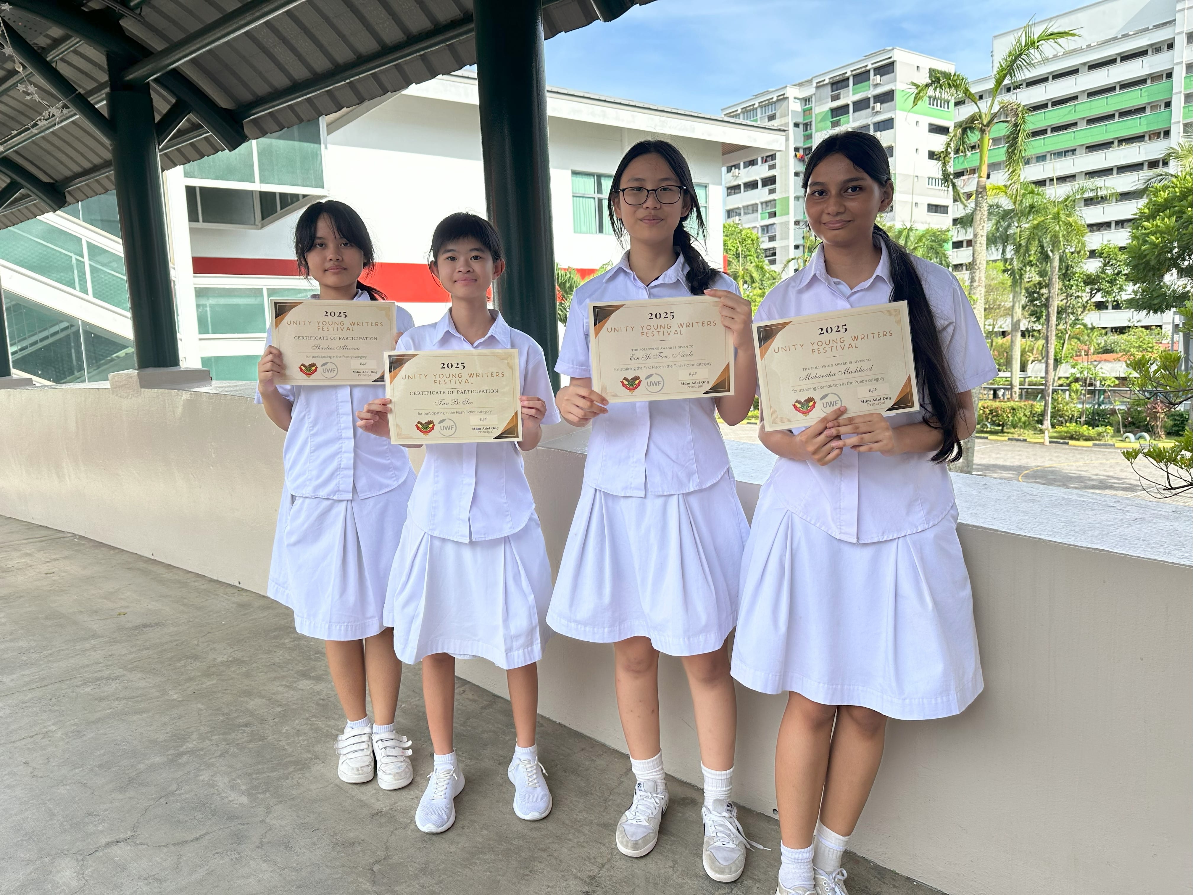 Four girls in white uniforms hold 2025 Unity Young Writers Festival certificates outdoors near school buildings.