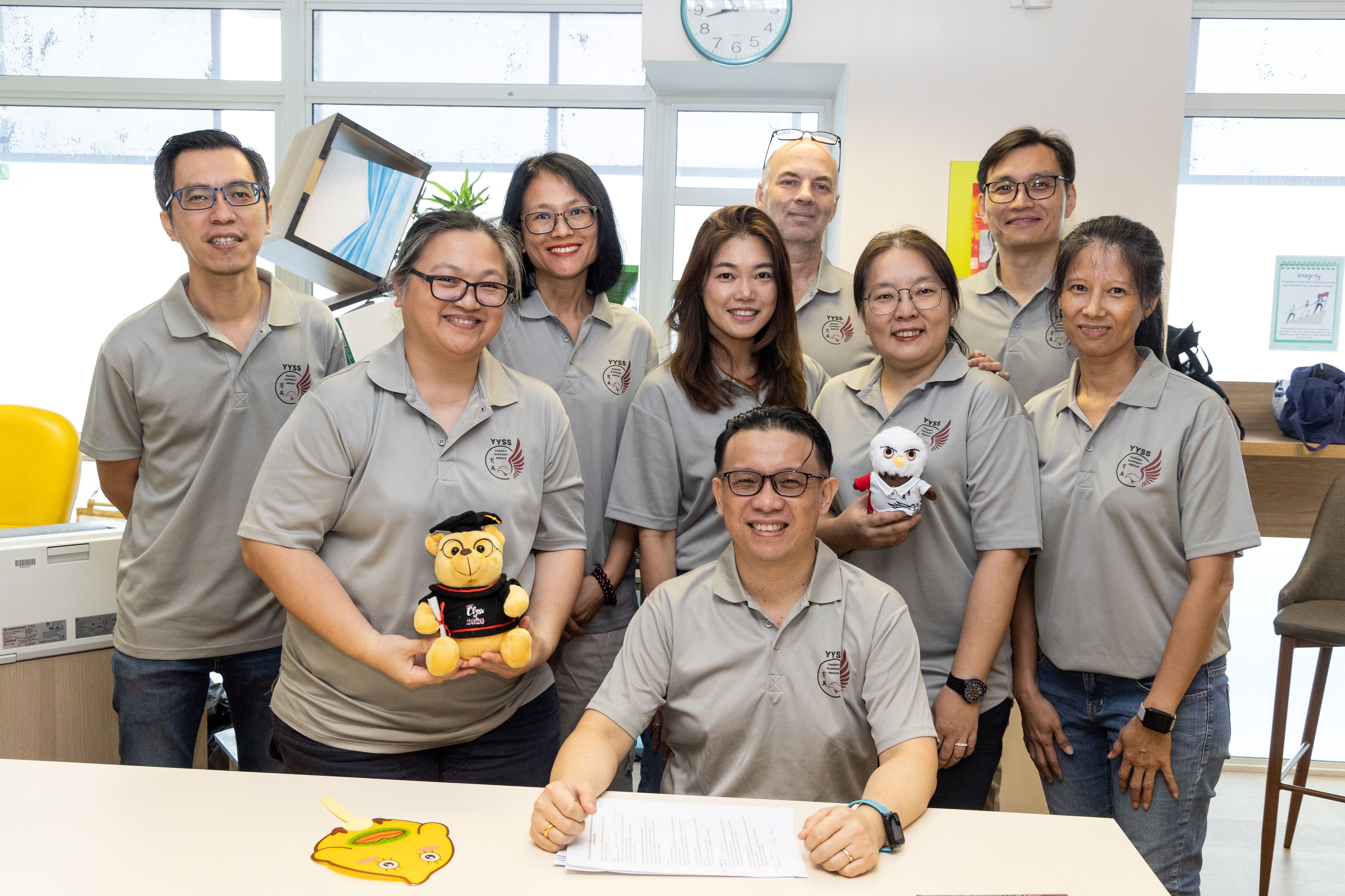 Nine people in matching grey YYSS polo shirts pose together indoors, two holding stuffed animal toys.