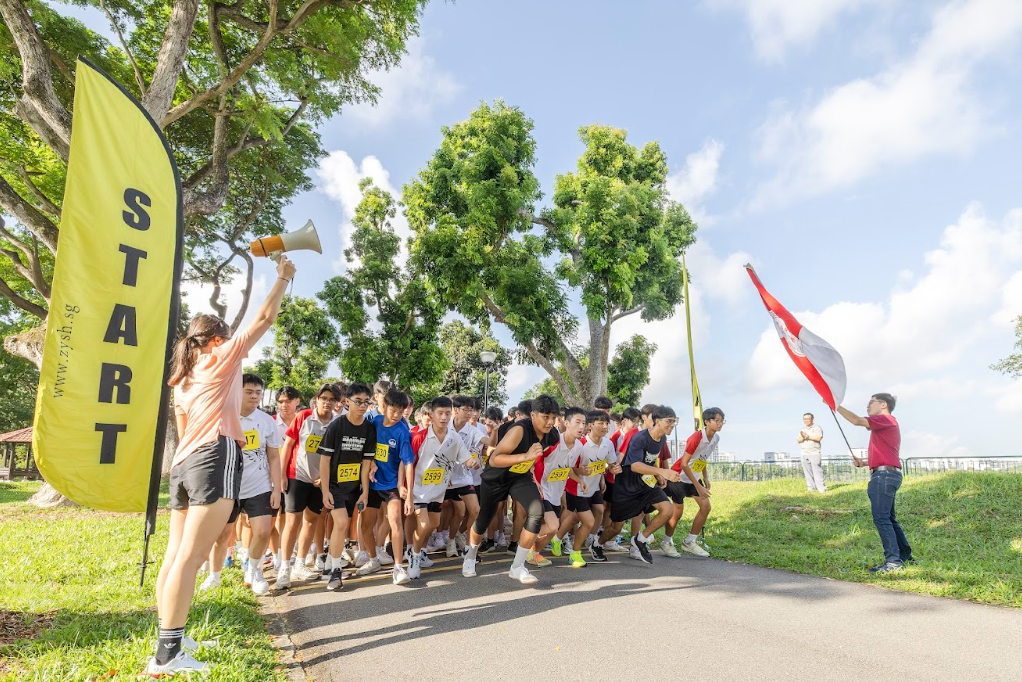 A group of young runners begin a race at a START banner as an official holds a megaphone outdoors.