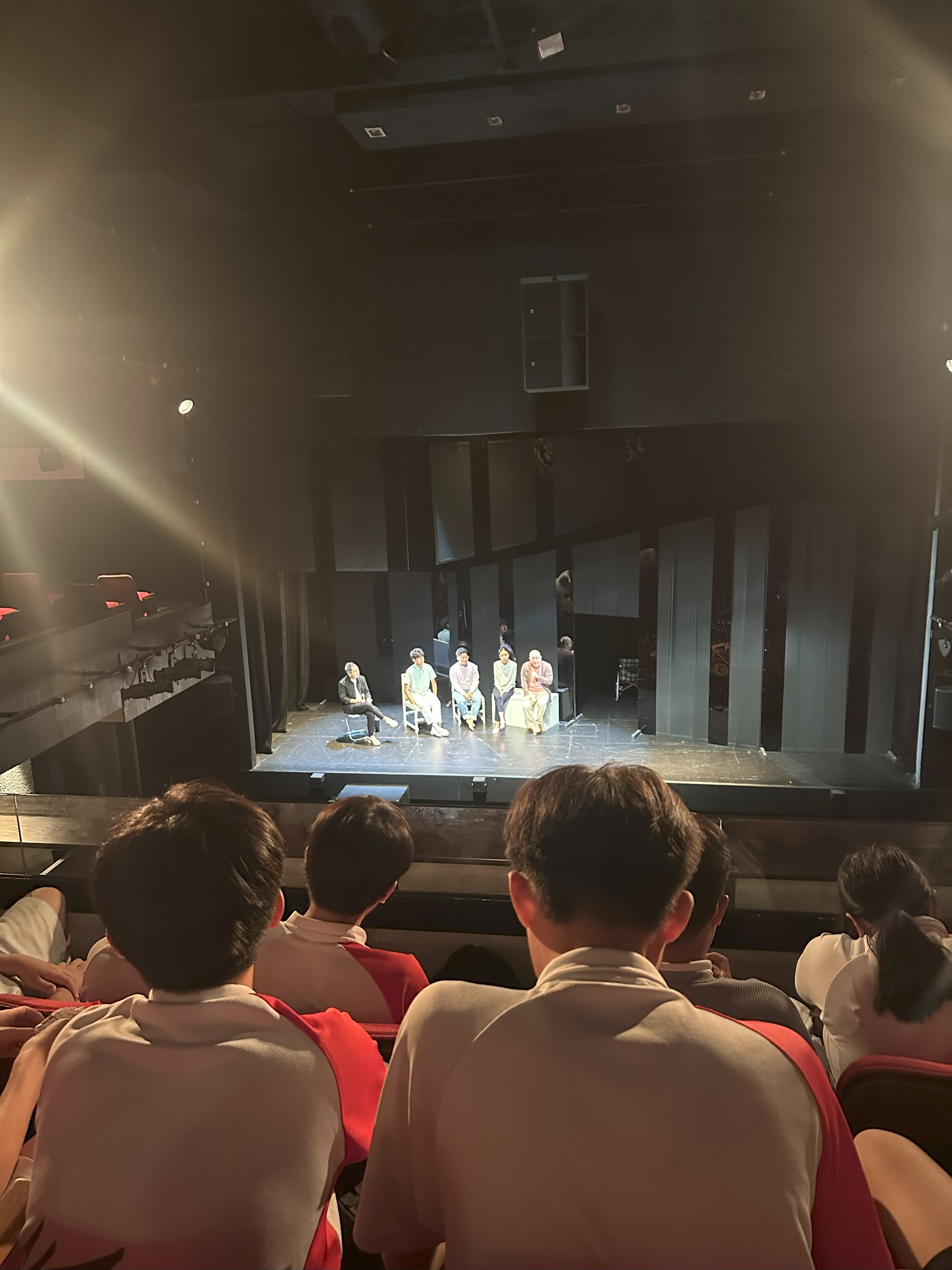Children in white and red uniforms sit in theater seats watching five people seated in chairs on a dimly lit stage.