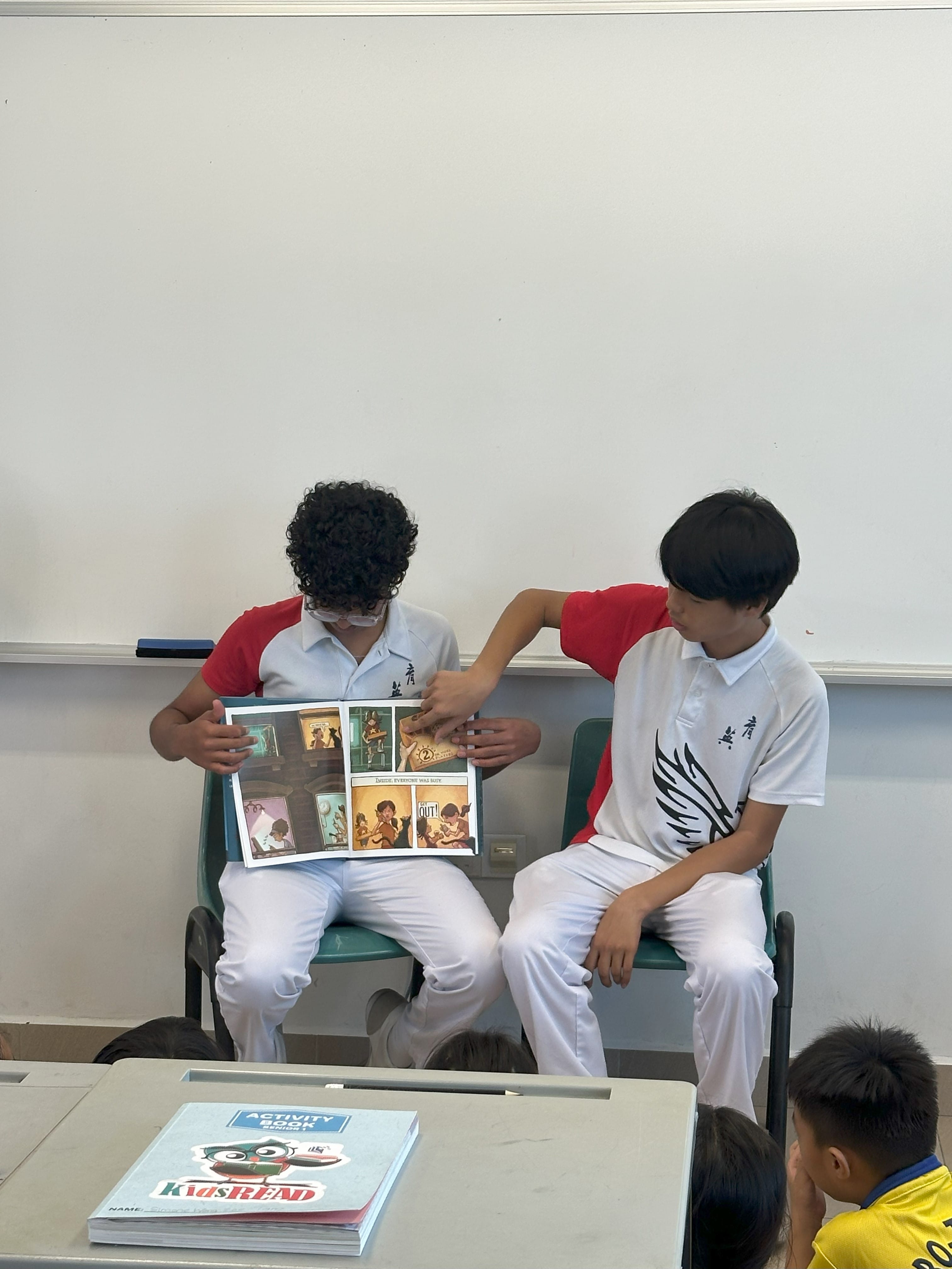 Two students in white uniforms sit in a classroom, one holding open a comic book while the other points at a page.