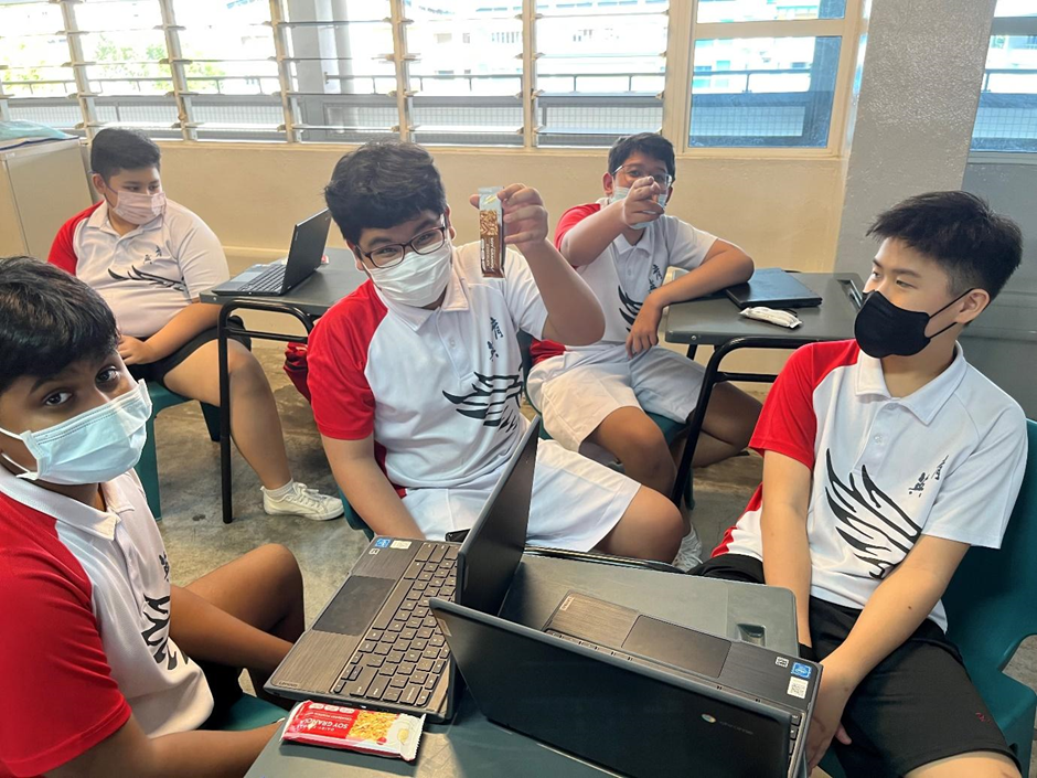 Five male students wearing face masks and matching uniforms sit at desks with laptops in a classroom.