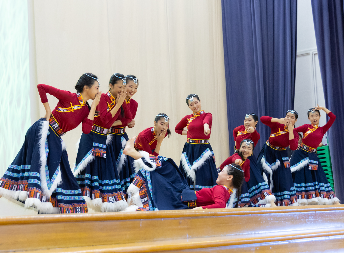 Group of young women in red tops and navy embroidered skirts with white fur trim performing a traditional dance on stage.