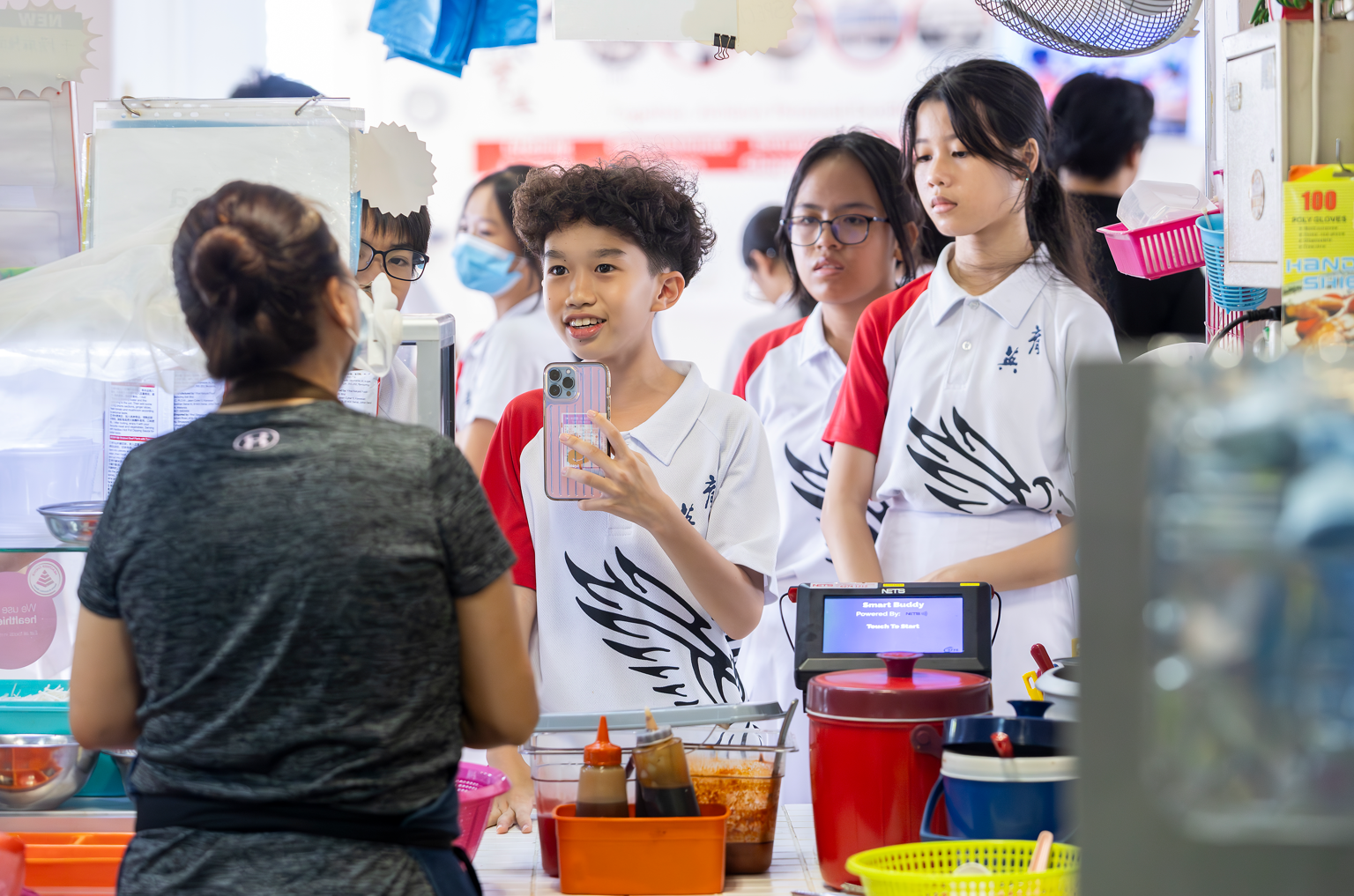 Students in white uniforms visit a food stall, one smiling boy holds a phone near a counter with condiments.