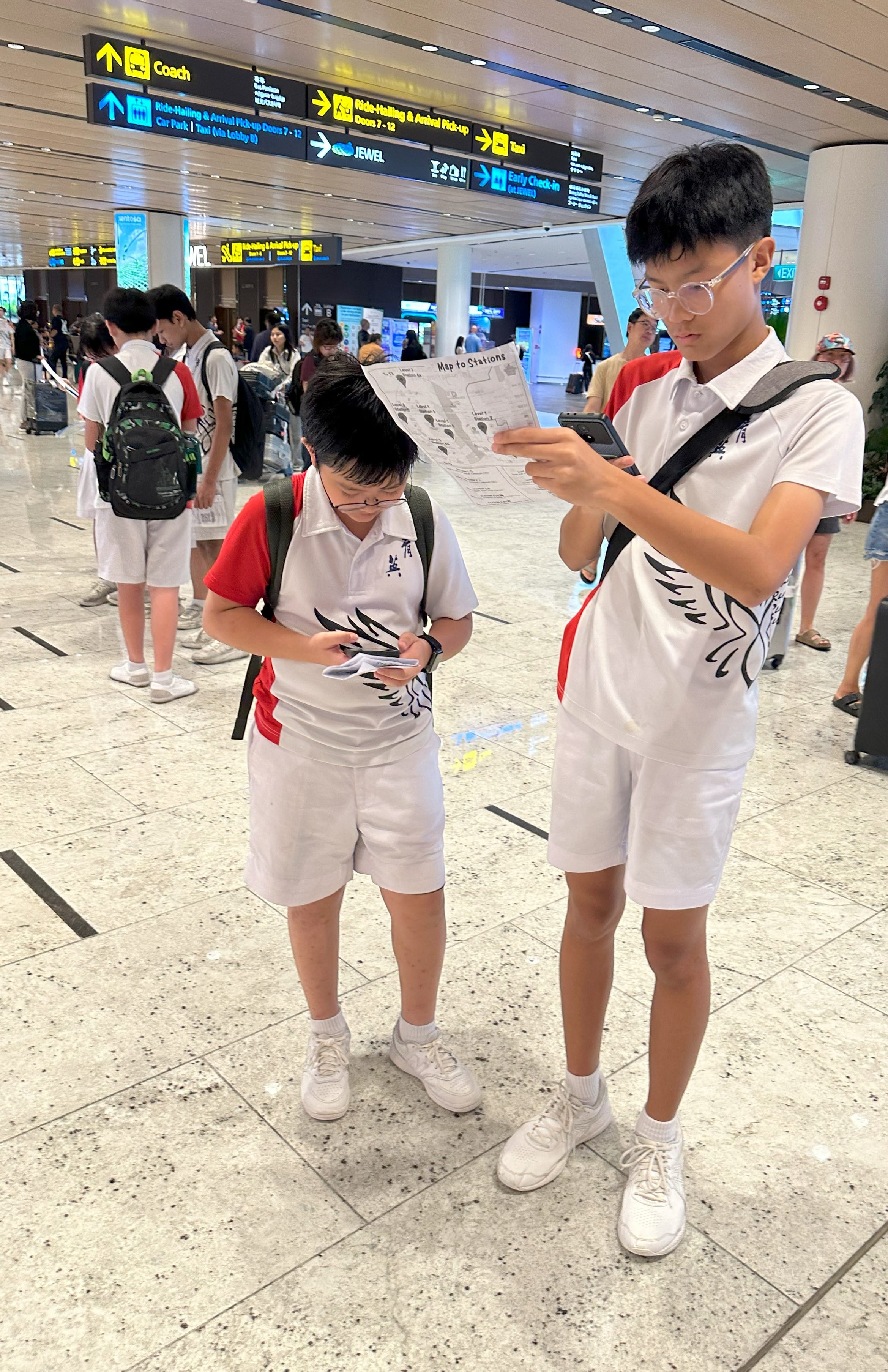Two boys in white school uniforms with backpacks study a map and phone inside a busy airport terminal.