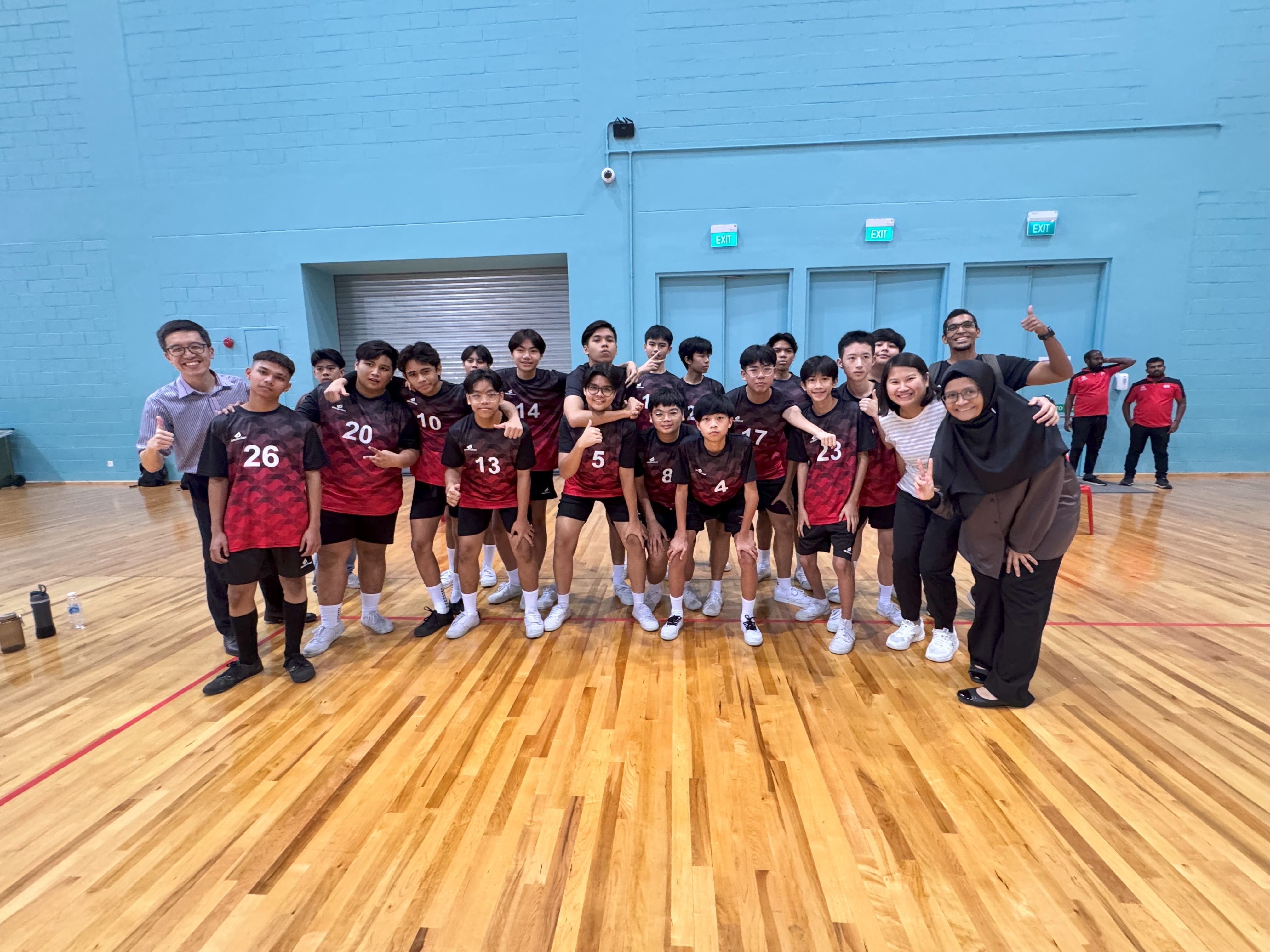 A youth volleyball team in red numbered jerseys poses with coaches on a wooden indoor court.
