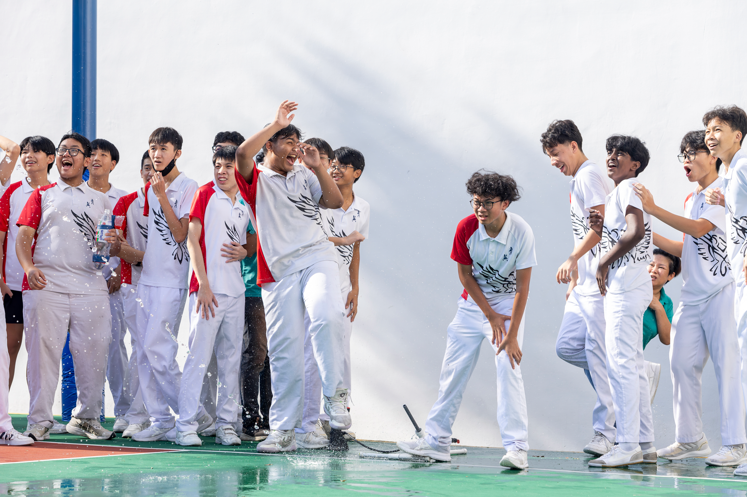 A group of teenage students in white uniforms laugh and cheer outdoors as water splashes on a green court.