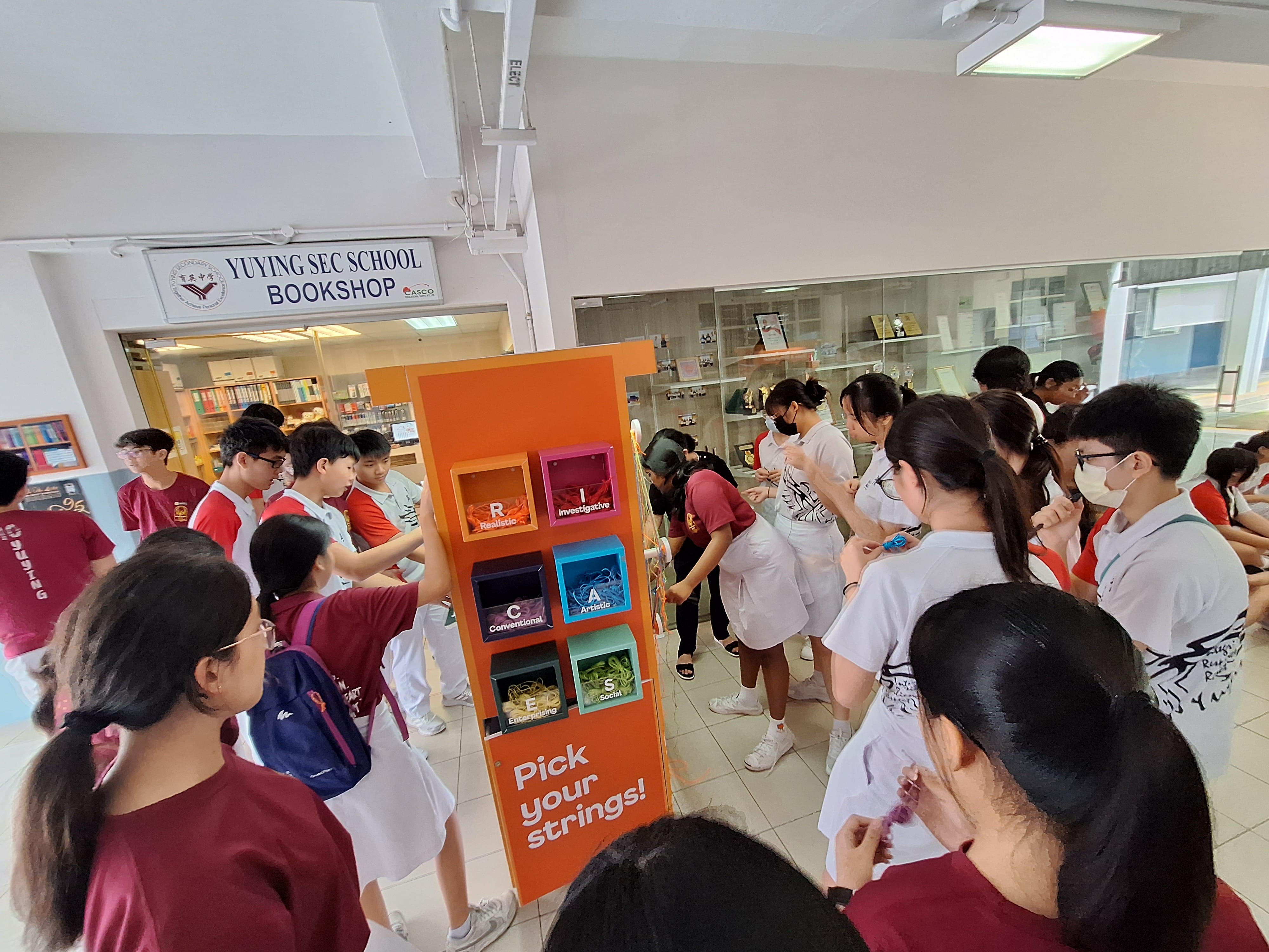 Students crowd around an orange "Pick your strings" RIASEC display board outside Yuying Sec School Bookshop.