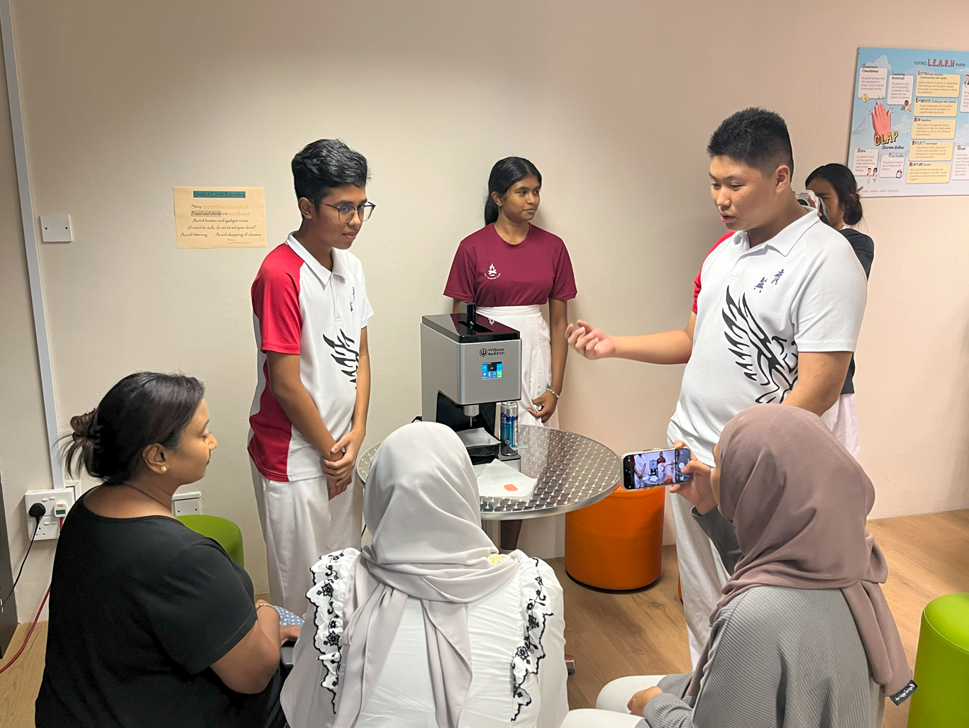 Students in school uniforms present a small machine to seated visitors in a classroom setting.