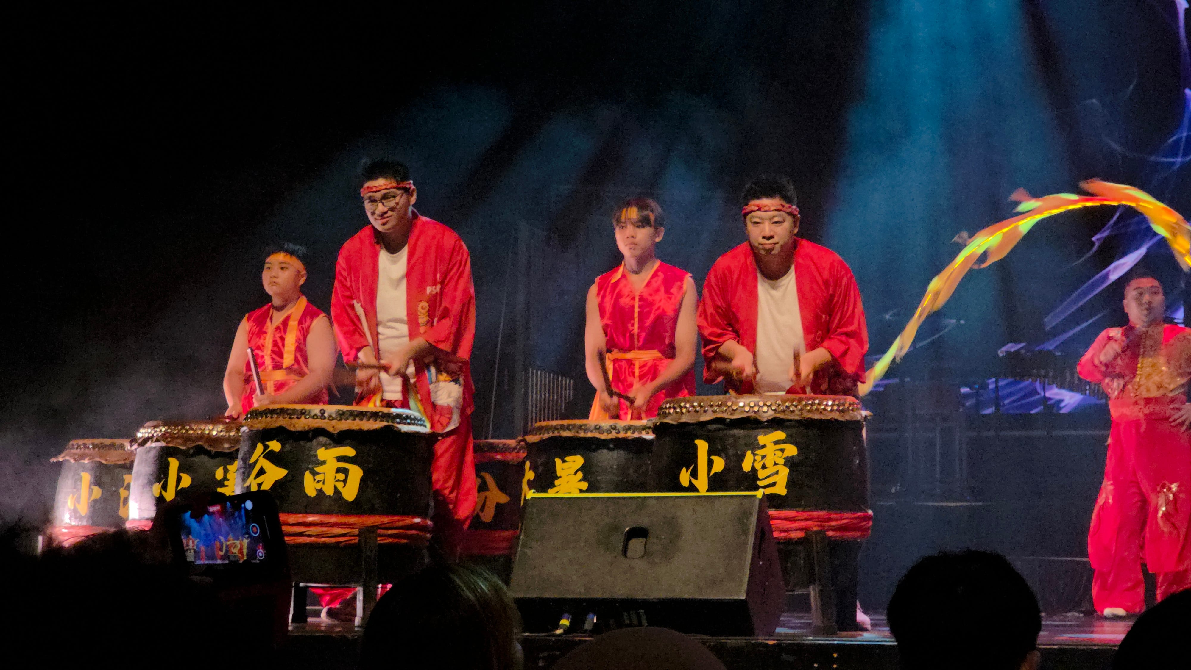 Four performers in red robes play large taiko drums with Chinese characters on a lit stage.