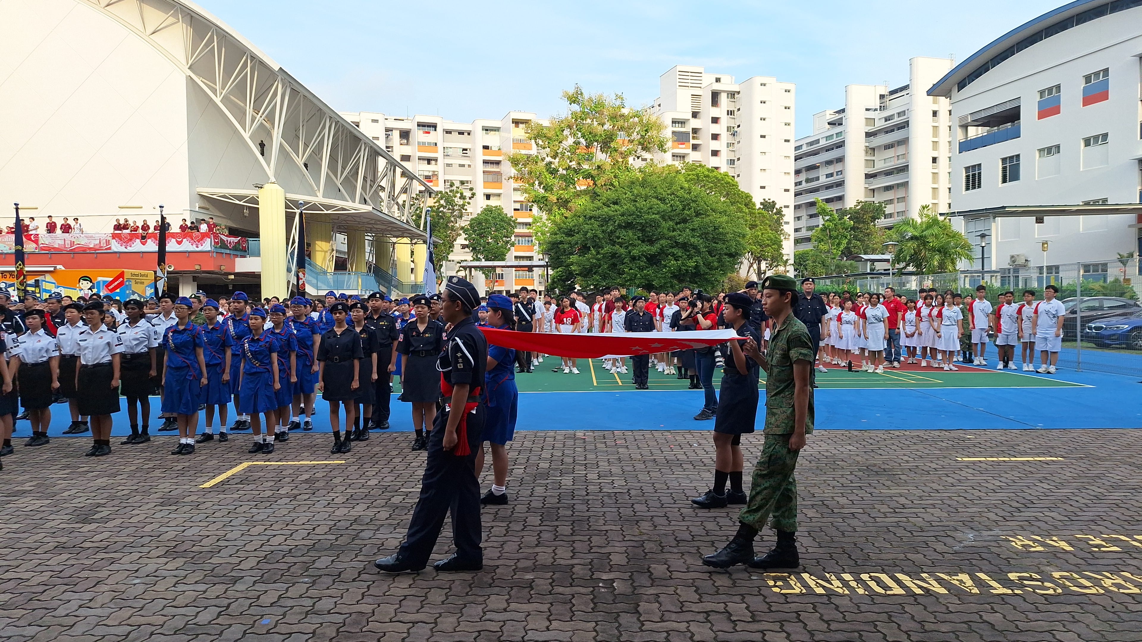 Students in uniforms stand at attention outdoors while a color guard carries a folded red flag during a school ceremony.