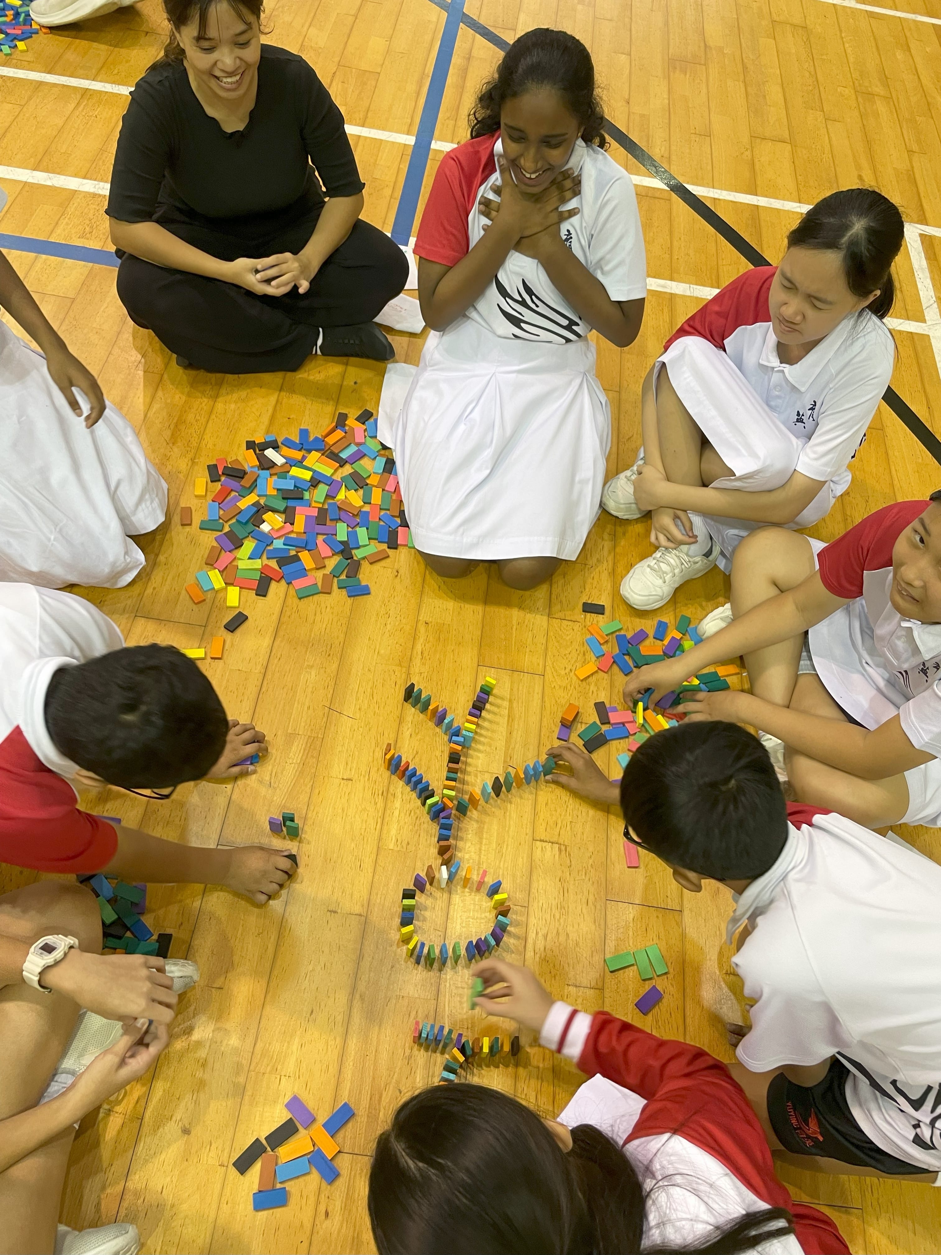Children and adults sit on a gym floor arranging colorful dominoes into a flower shape, smiling and engaged.