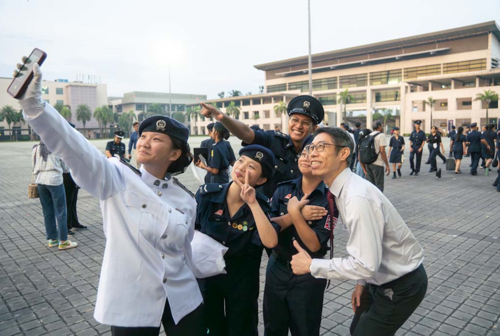 Five people in uniforms and formal wear smile and pose for a selfie outdoors near a large building.