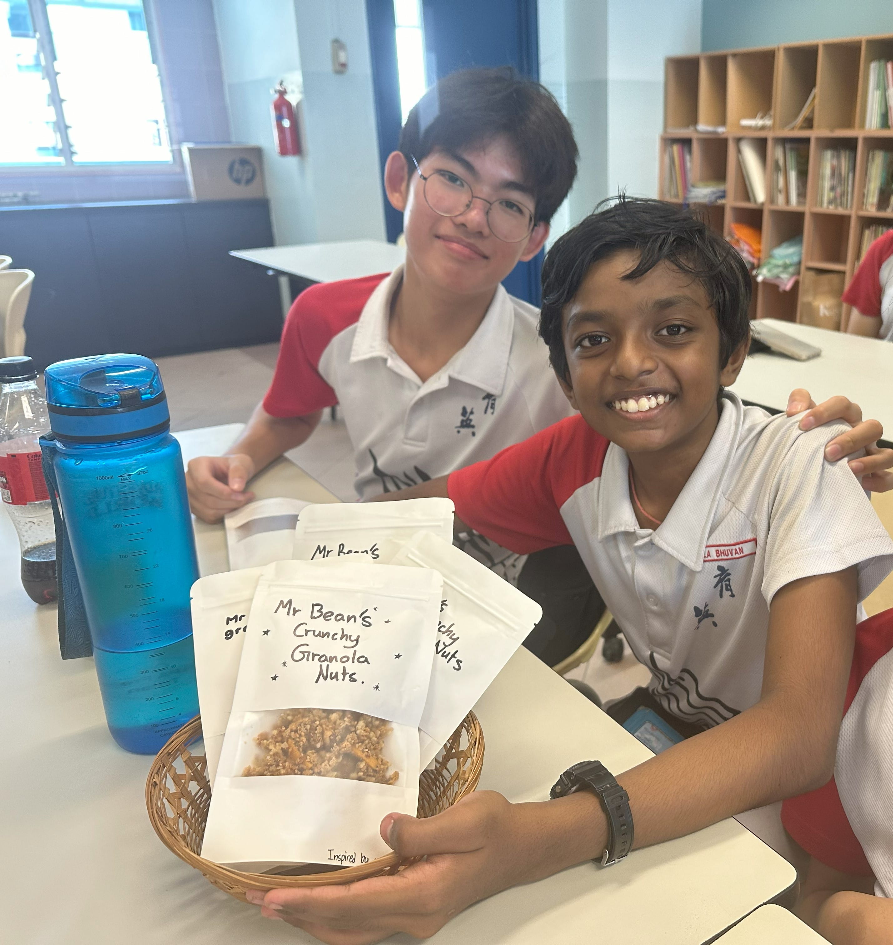 Two smiling school boys holding a basket with packets of Mr Bean's Crunchy Granola Nuts in a classroom.