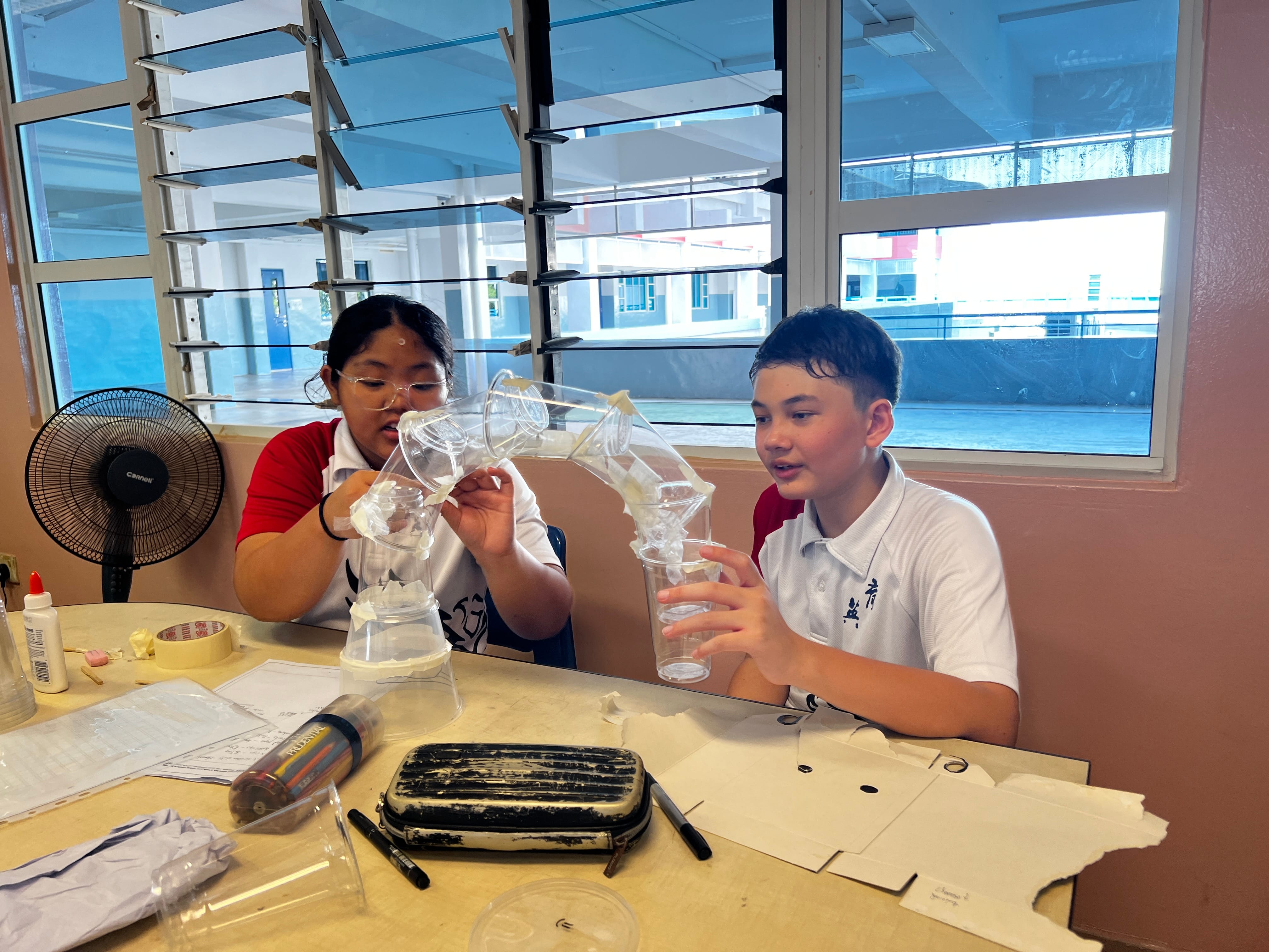 Two students in school uniforms build a structure using clear plastic cups taped together at a classroom desk.
