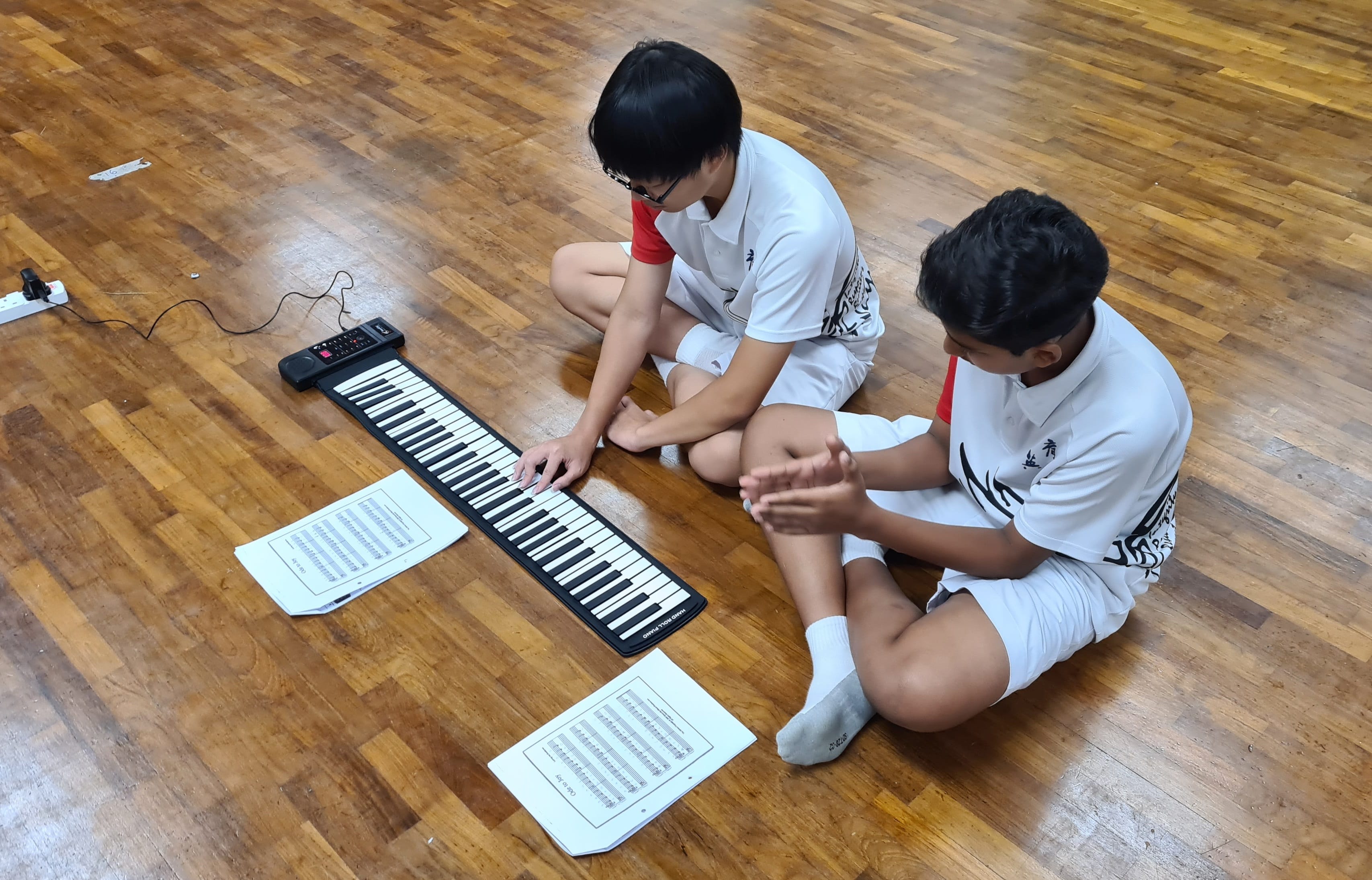Two students in white shirts sit on a wooden floor playing a roll-up flexible piano keyboard with sheet music nearby.
