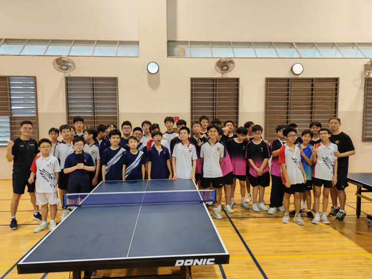 A large group of young male students and coaches pose together behind a Donic table tennis table in an indoor gym.