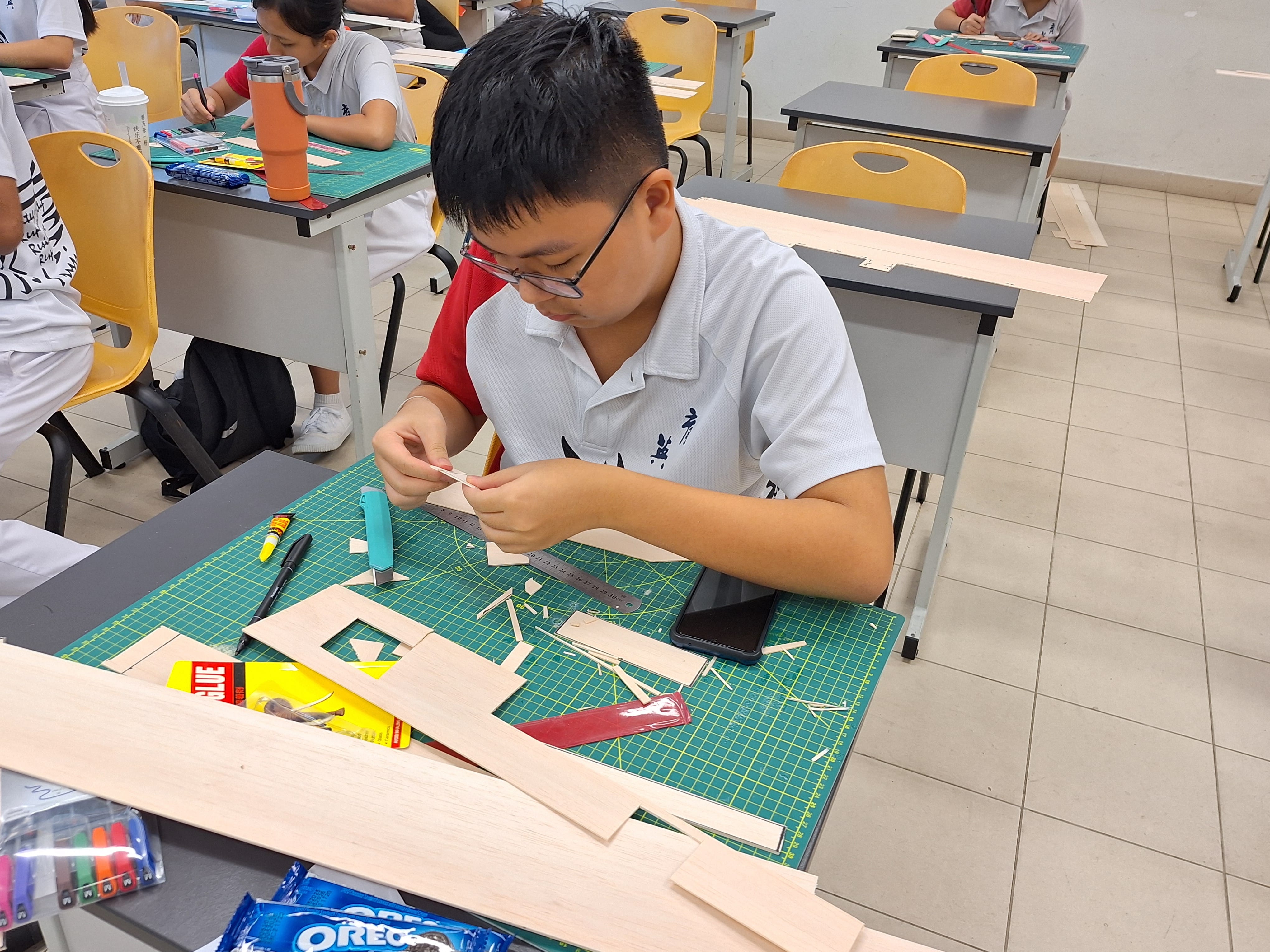 A young student wearing glasses assembles a wooden model on a green cutting mat in a classroom.