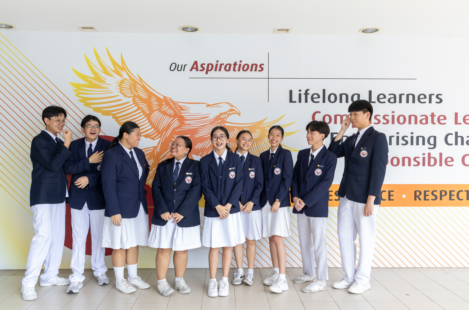 Nine students in navy blazers and white uniforms stand laughing before a school wall reading Our Aspirations.