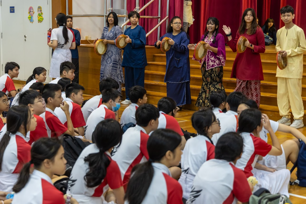 Students in traditional attire play frame drums before a seated audience of uniformed schoolchildren in a hall.
